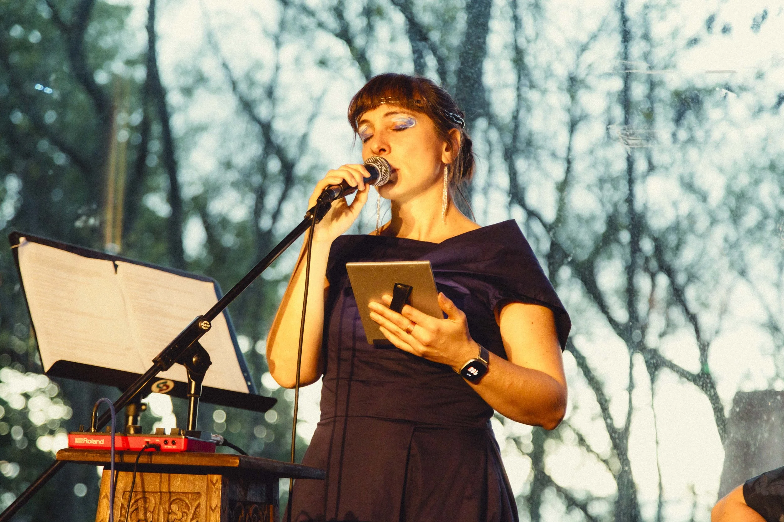 A woman in a dress with blue eyeshadow is singing into a microphone while standing outdoors. There is electronic music equipment on a small wooden table, and trees are visible in the background.