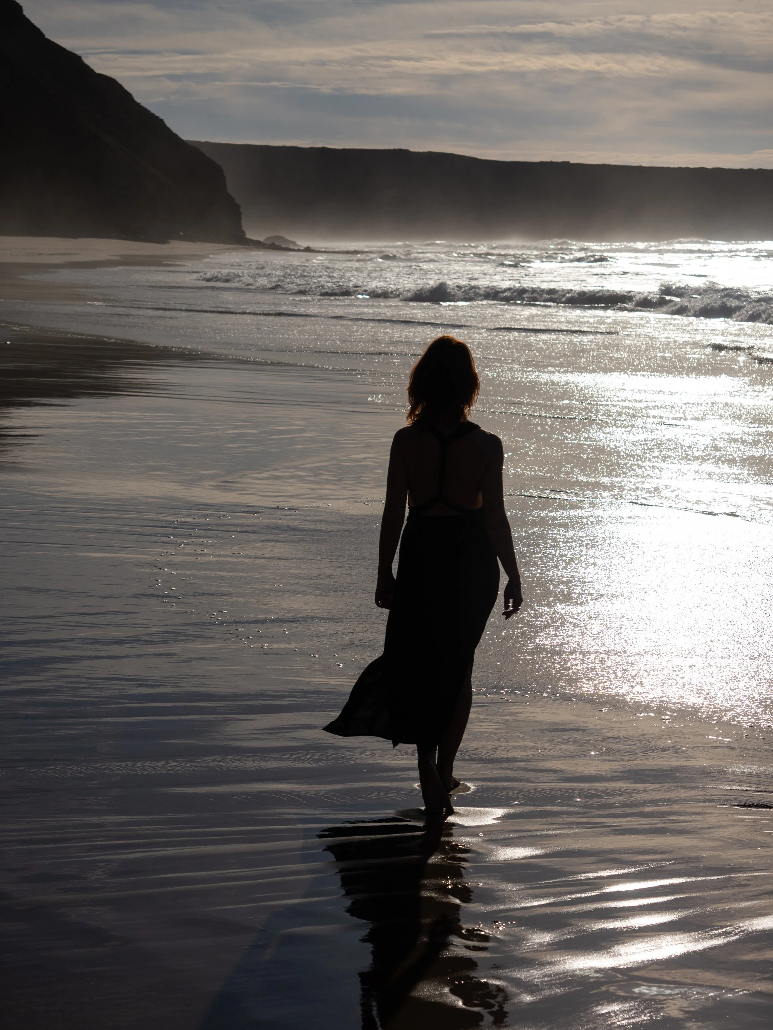 Femme silhouette marchant sur la plage au coucher du soleil, reflet sur le sable humide, mer agitée et falaises en arrière-plan.