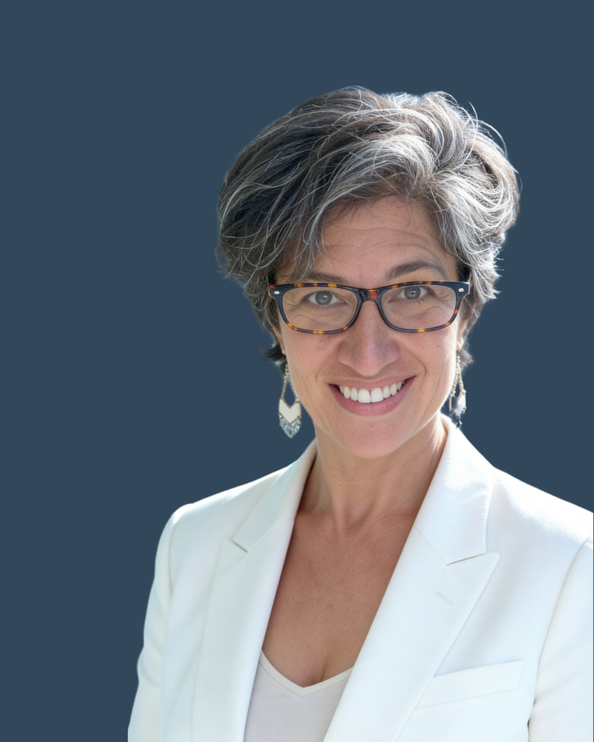A smiling woman with short gray hair, glasses, and earrings, wearing a white blazer, against a dark blue background.