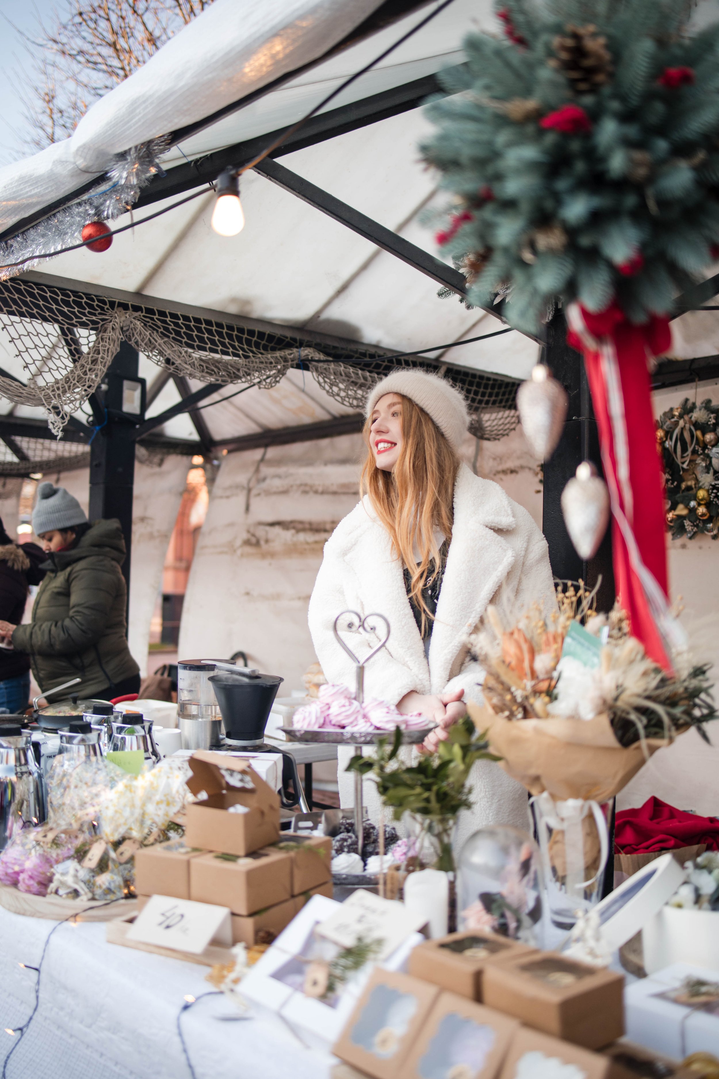 A woman dressed in a white coat and beige beanie smiling at a holiday market stall decorated with Christmas ornaments and greenery.