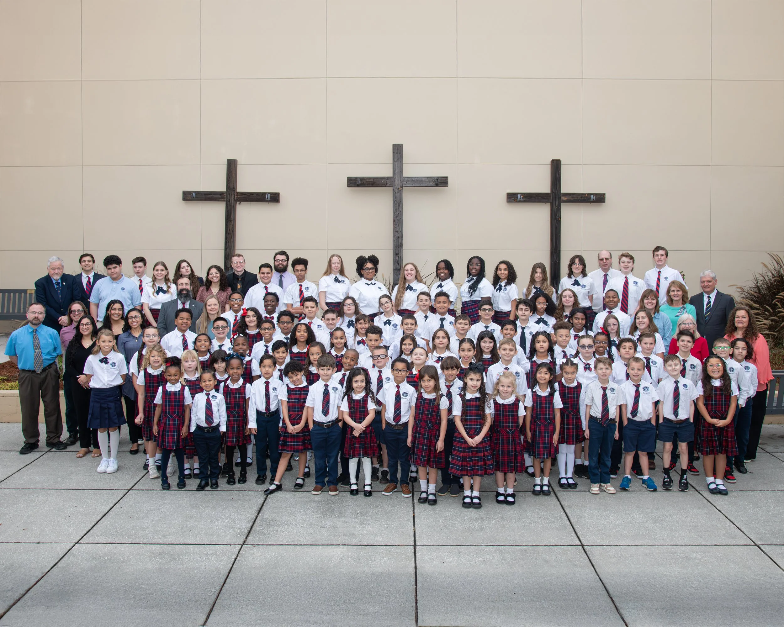 A large group of elementary school students and teachers posing outdoors in front of a wall with three large wooden crosses, likely a school photo taken during religious education day.