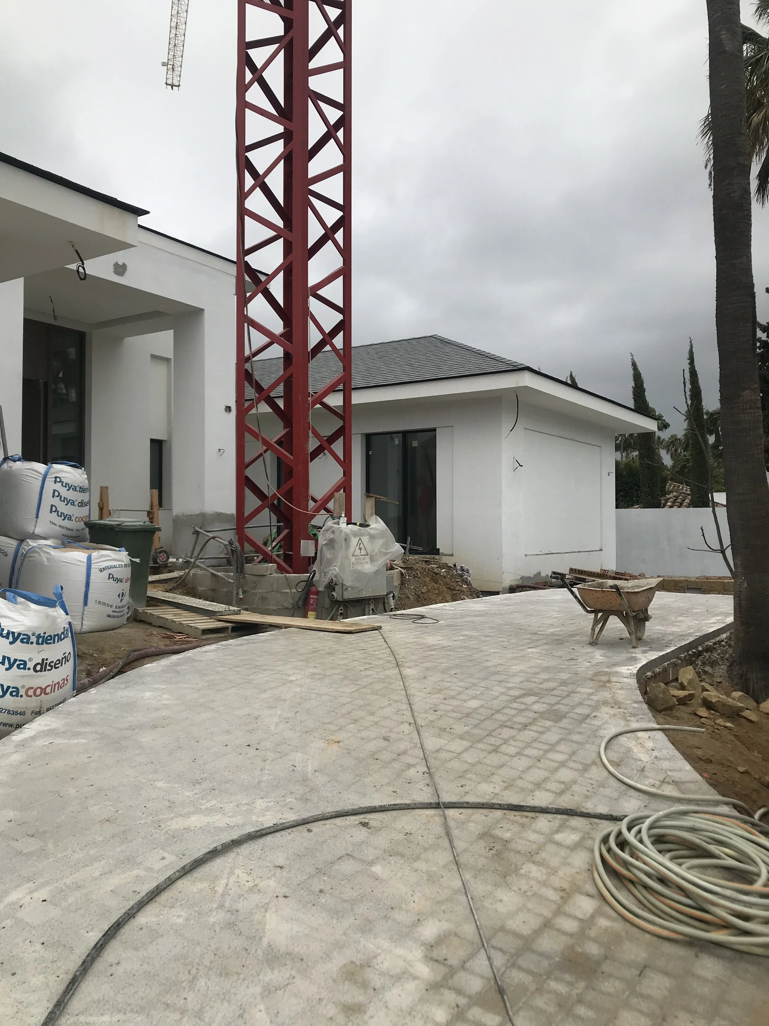 Construction site with a white modern house, a red construction crane, and building materials including bags and a wheelbarrow. Overcast sky with palm trees in the background.