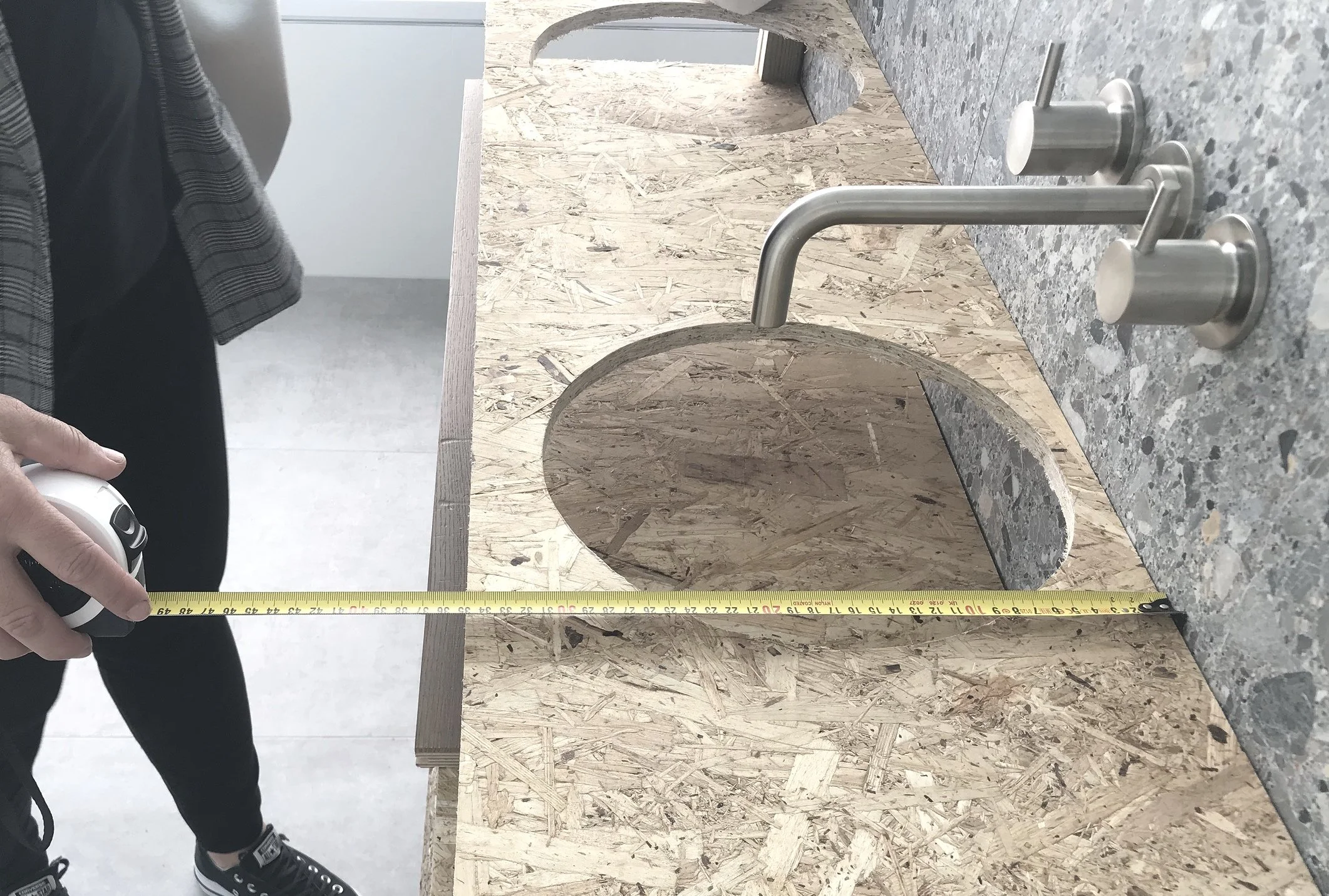A person measuring a piece of particle board with a tape measure, with a granite countertop and stream of water in the background.