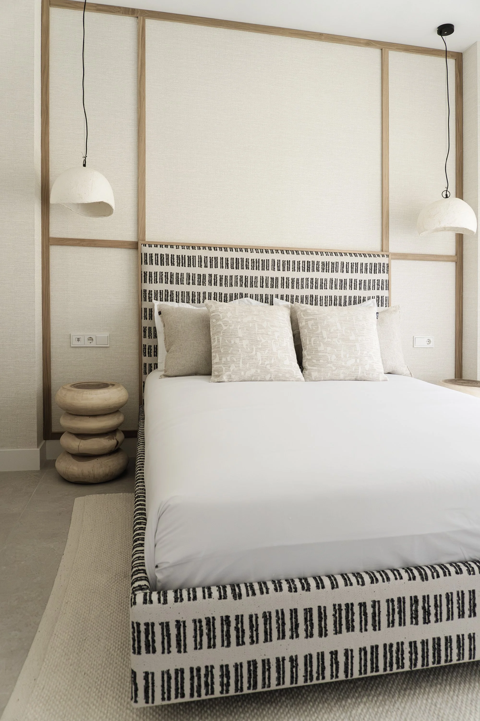 A modern bedroom with a bed, decorative headboard, beige and striped pillows, wooden side tables, fabric nightstand, and hanging pendant lights in a neutral color palette.