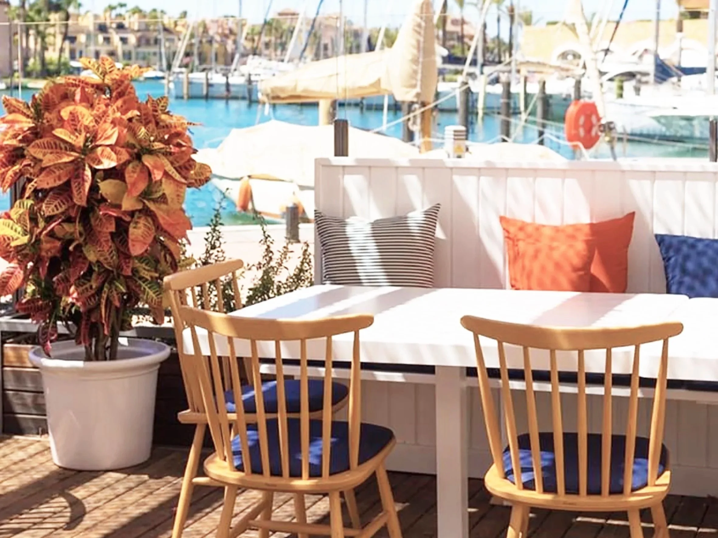 Outdoor patio seating area overlooking a marina with sailboats, featuring wooden chairs, a round table, and decorative pillows.