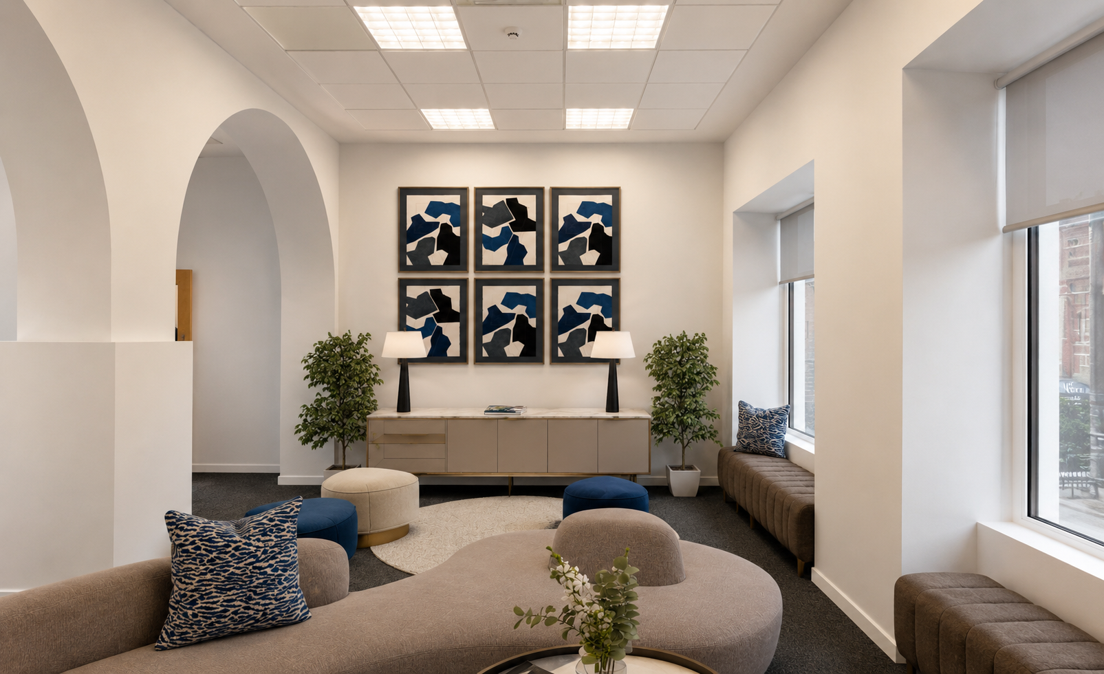 Office reception area with arched entryways, abeige sofa with blue and beige pillows, round beige and blue ottomans, sideboard with lamps, framed abstract artwork, potted green plants, window seats and large windows with white blinds.