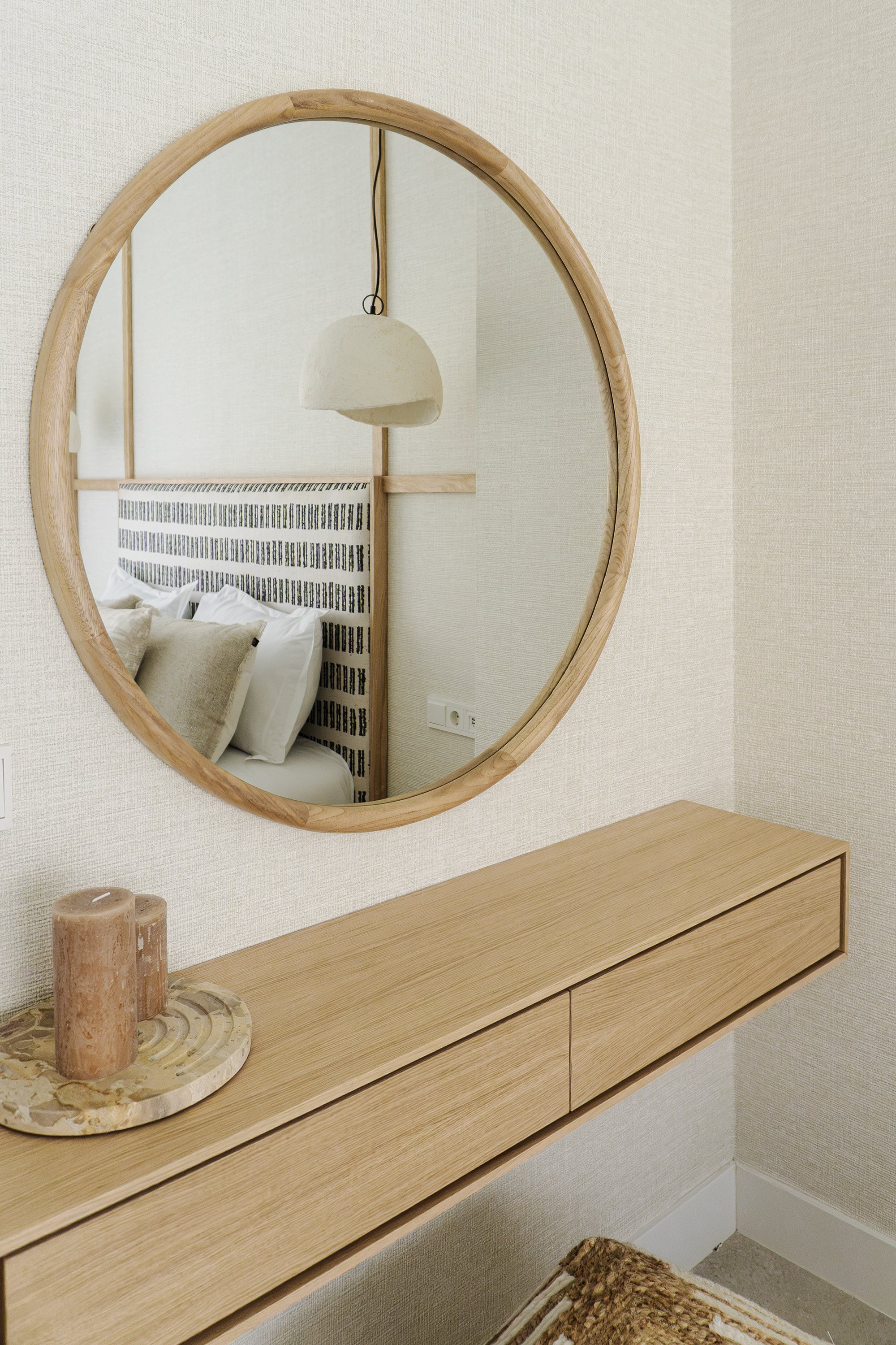 A wooden dresser beneath a round mirror with a light wood frame. On the dresser, there is a round base with a textured candle holder and two candles inside. Part of a bed with a headboard and pillows is reflected in the mirror, and a wall with a powe