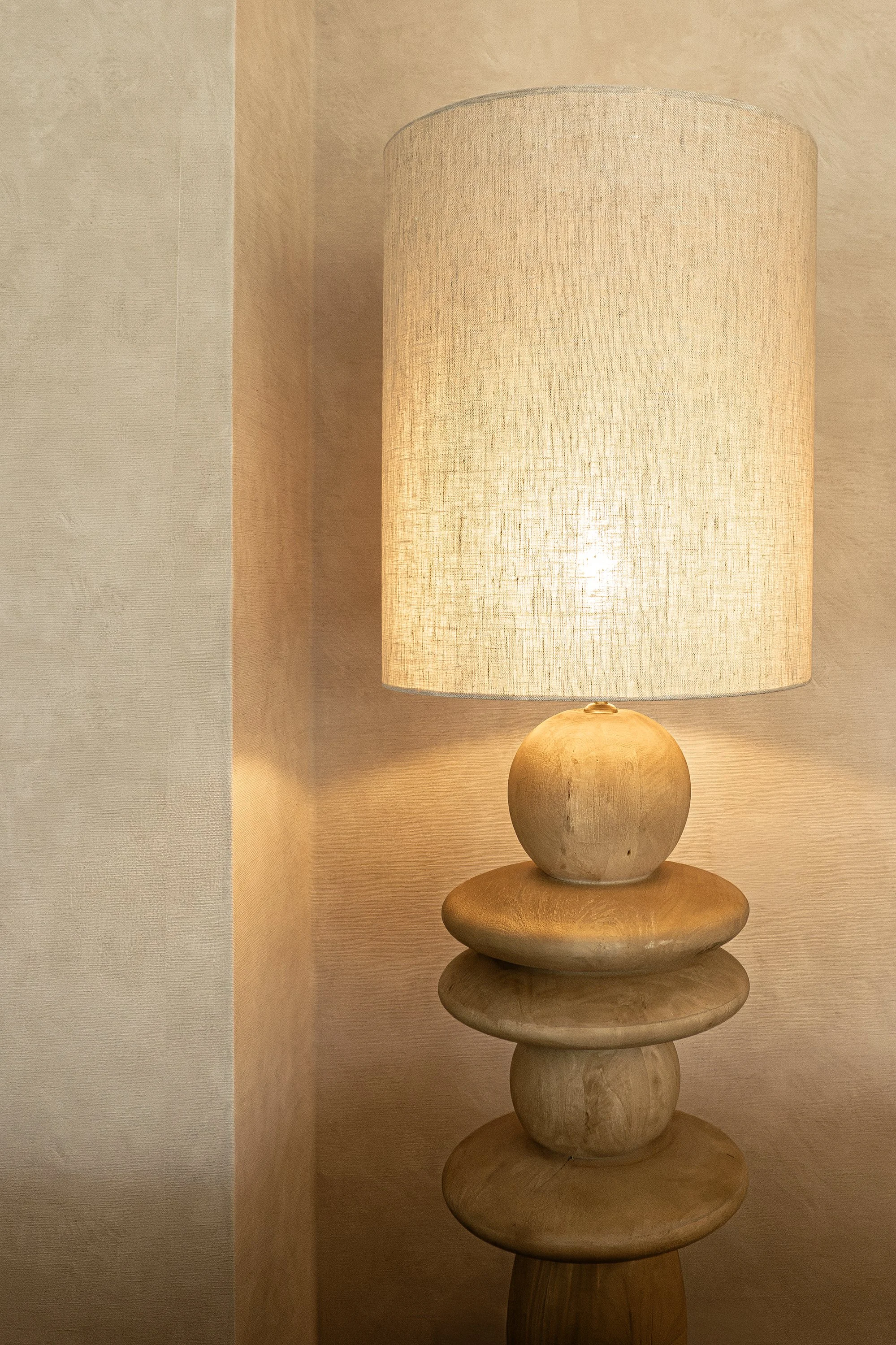 A wooden table lamp with a beige fabric lampshade, placed on a multi-tiered wooden base, situated against a beige wall.