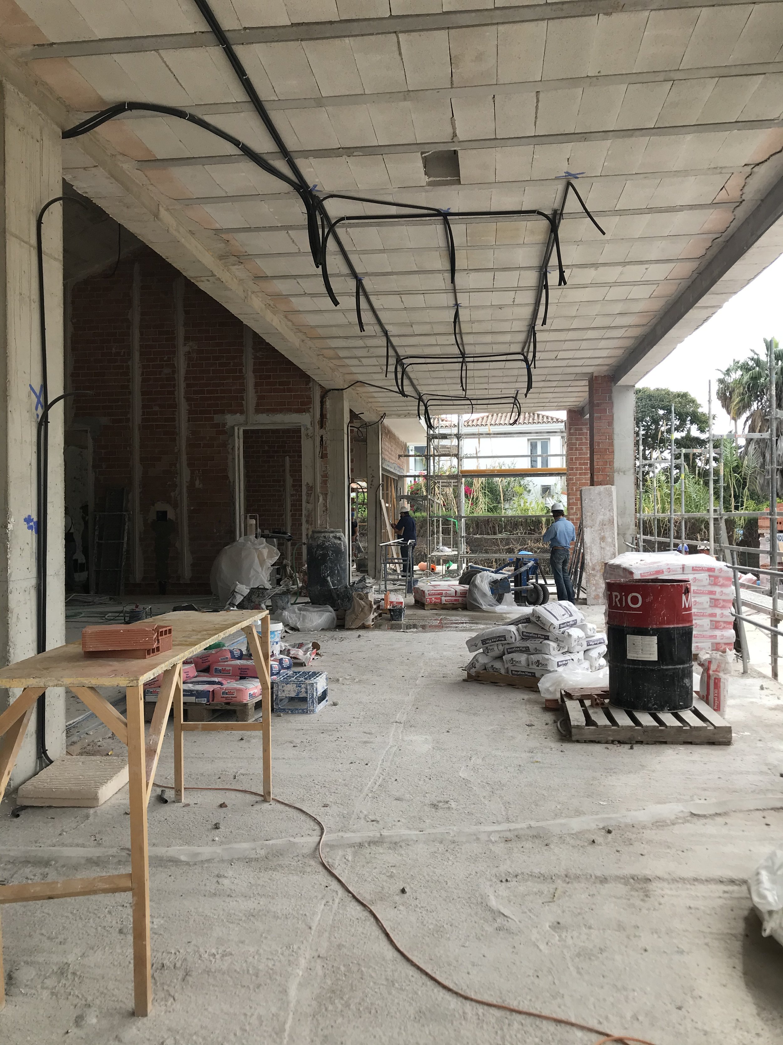 Construction site with workers, scaffolding, building materials, electrical wiring on ceiling, and partially built walls.