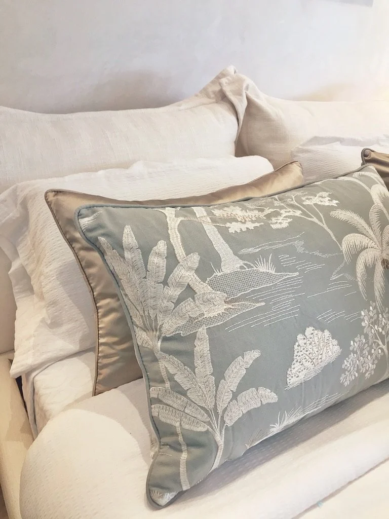Decorative pillows on a white sofa, with the front pillow featuring an embroidered nature scene with trees, plants, and a lake in light colors.