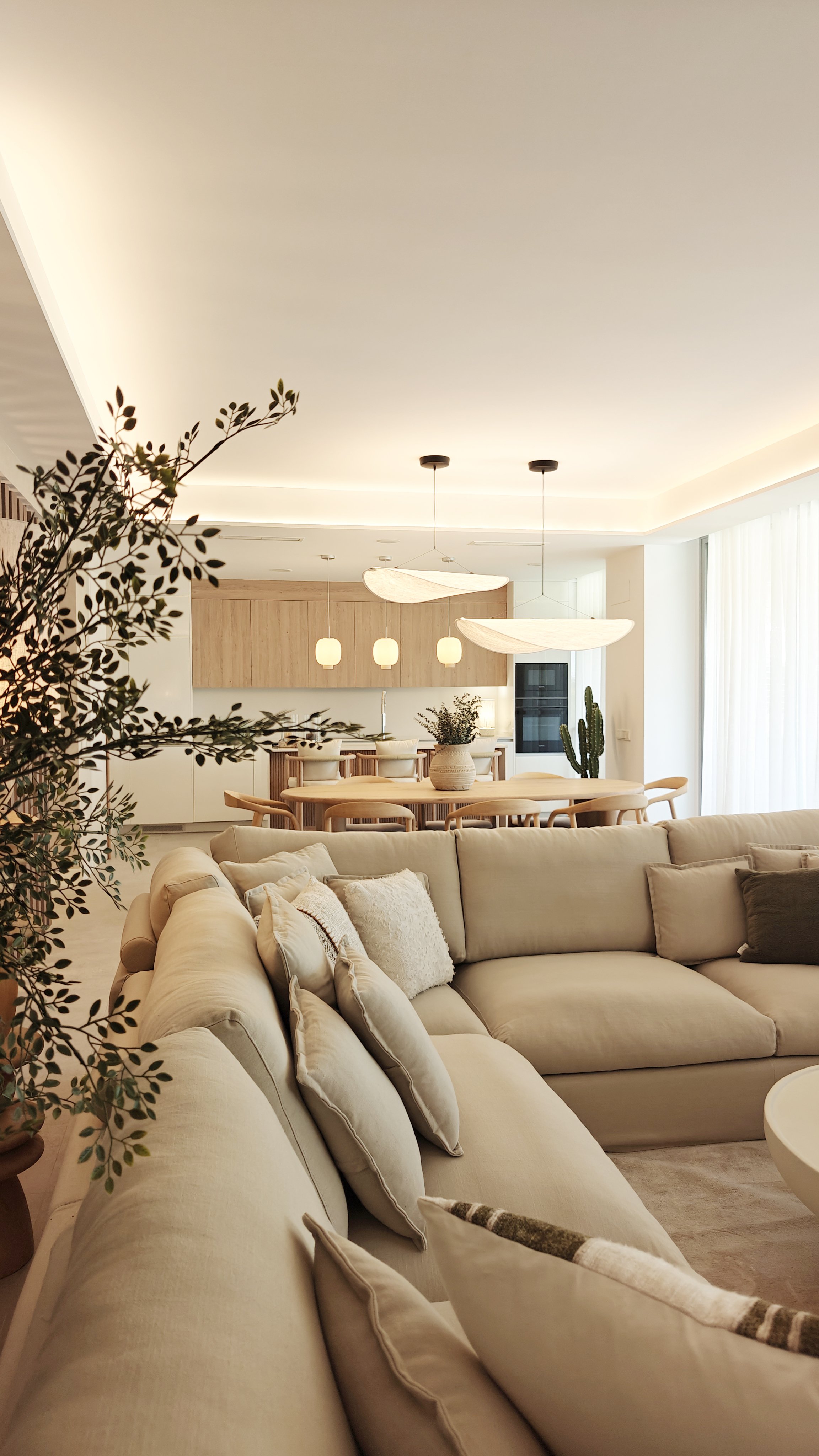 Living room with beige sectional sofa, throw pillows, potted plant, modern white pendant lights, wooden dining table with chairs, and large window with sheer curtains.