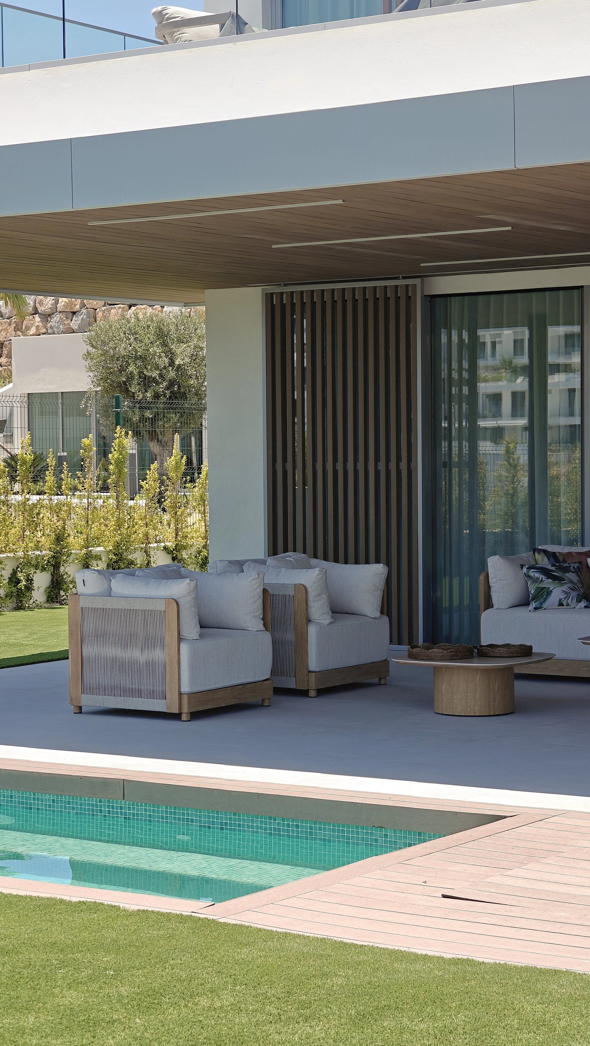 Outdoor patio area with white cushioned seating, a wooden coffee table with trays, a pool with turquoise tiles, a green lawn, and a modern building with large windows and wooden accents.