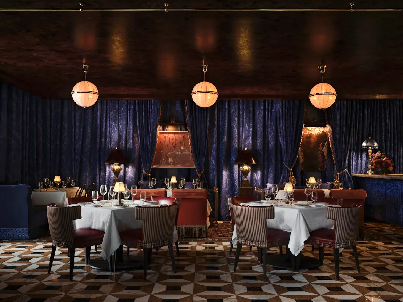 Elegant restaurant dining room with round tables covered in white tablecloths, set with wine glasses and silverware. The room has dark blue curtains, vintage lamps, and a checkered wooden floor.