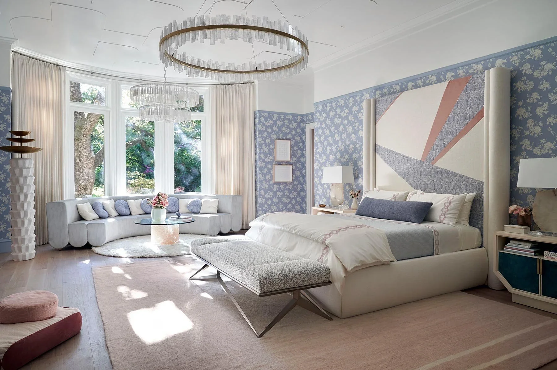 Bright bedroom with large window, blue floral wallpaper, white bed with a large headboard, a bench at the foot of the bed, a sitting area with a curved sofa, a round coffee table with flowers, and modern lighting fixtures.