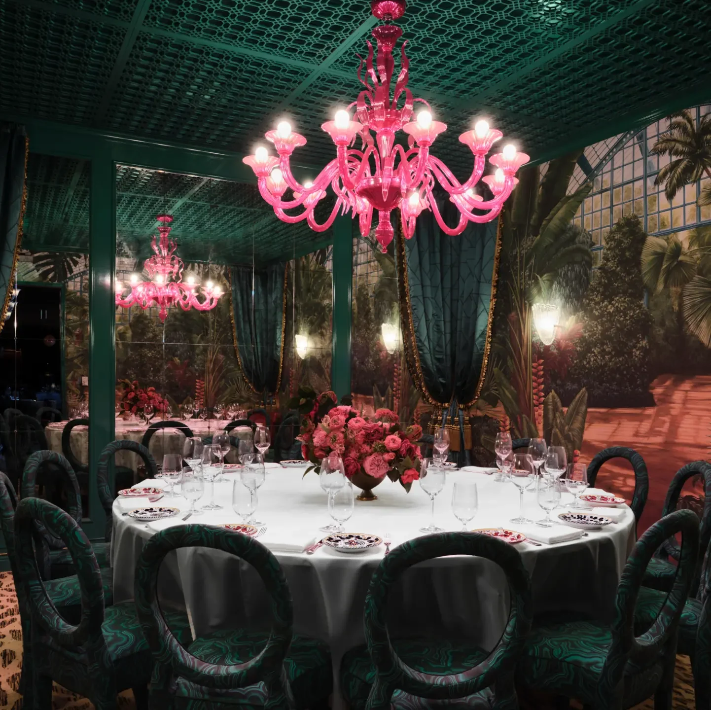 Elegant dining room with a round table set for a formal meal, decorated with a large pink floral centerpiece, wine glasses, and ornate plates. The room features dark teal curtains with gold trim, tropical wall murals, and pink chandeliers hanging from a patterned ceiling, creating a luxurious ambiance.