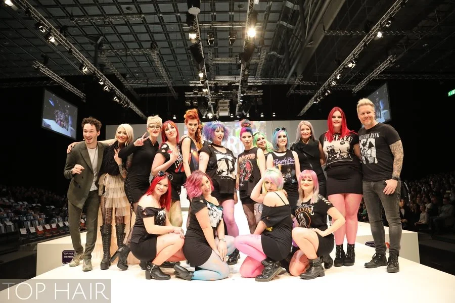 Group of diverse women and two men on a fashion runway stage during a hair or fashion show with bright lighting, audience in background, and a large screen displaying the event