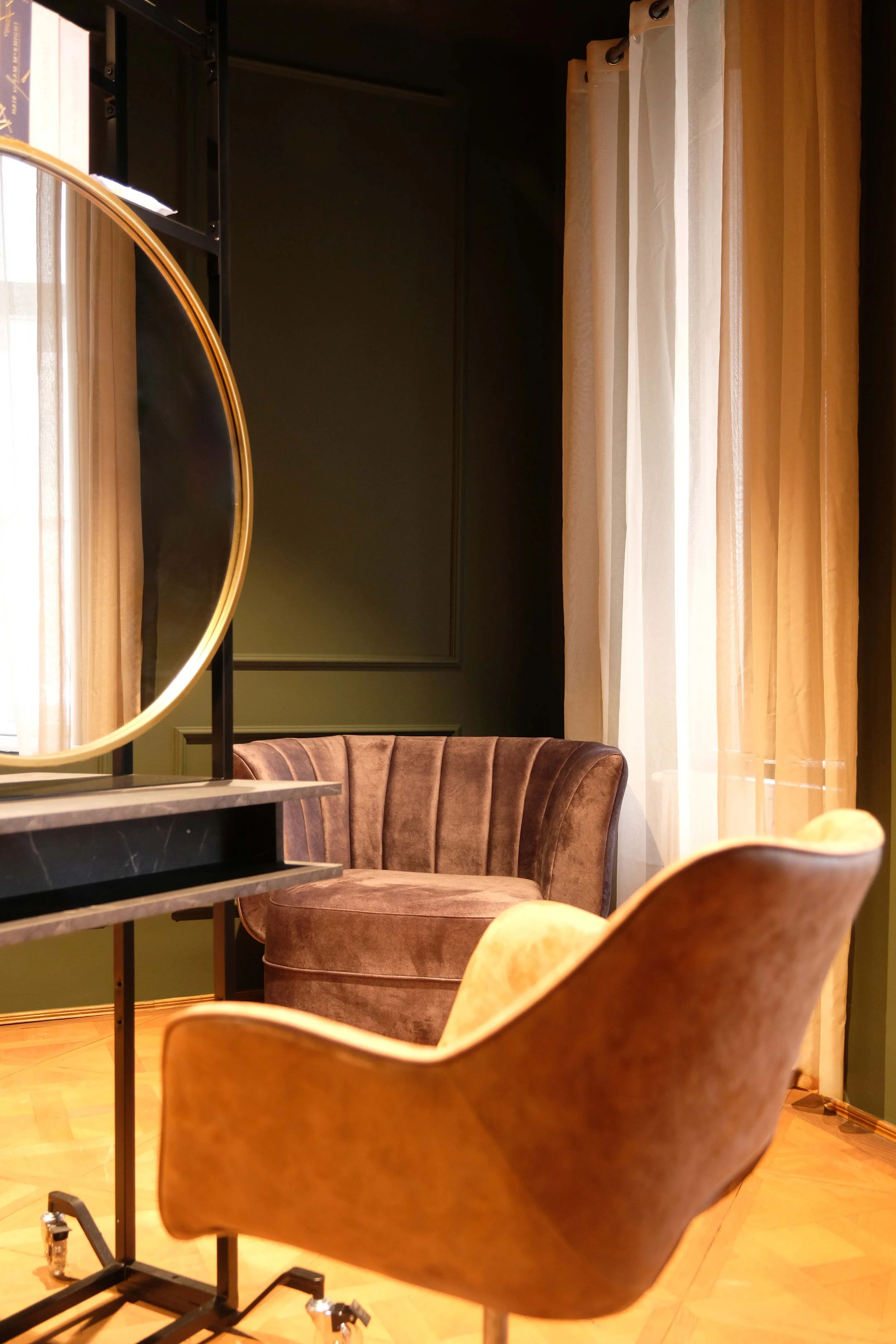 Cozy corner with brown velvet armchair and yellow fabric chair near a window with white and beige curtains, and a black shelf with a round mirror.