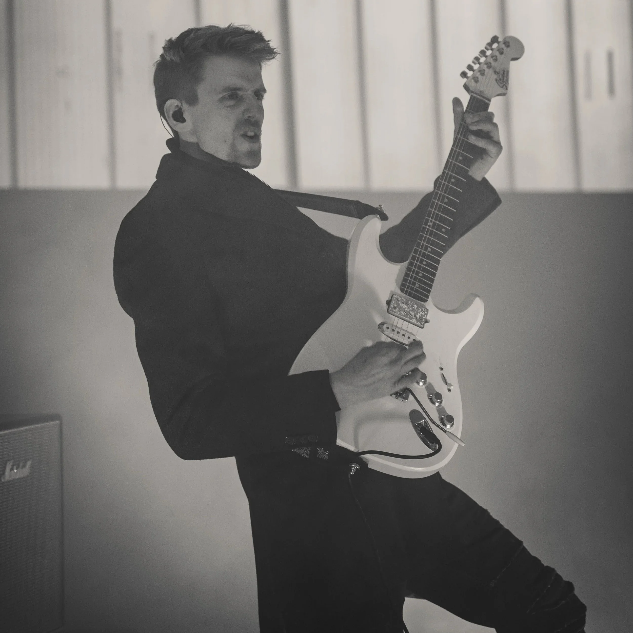 A young man playing an electric guitar on stage, wearing a black jacket and ripped jeans, with a focused expression.
