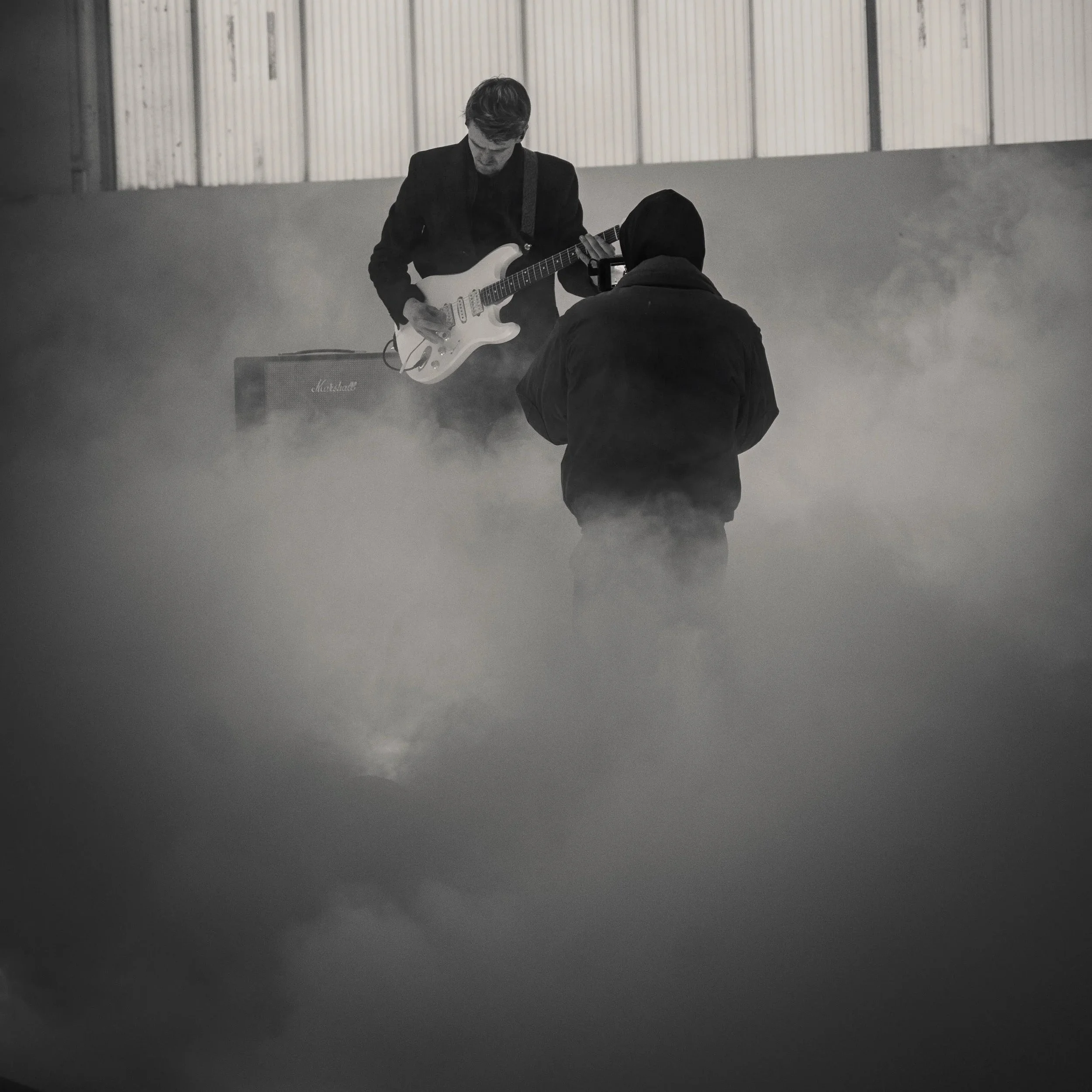 A person in a black hoodie is filming a guitarist playing an electric guitar in a foggy or smoky environment.