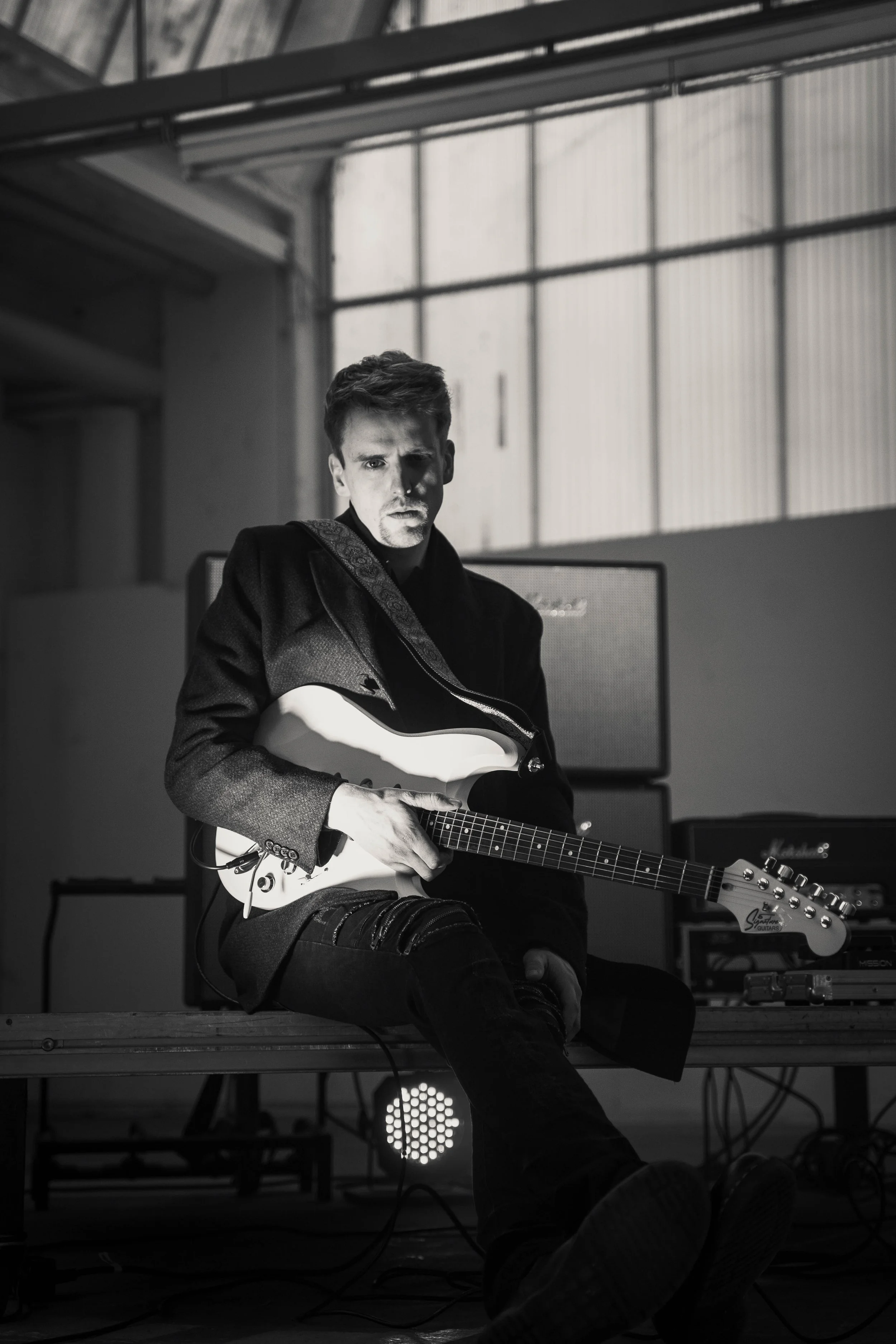 A black and white photo of a young man sitting on a wooden platform with an electric guitar, in an industrial-style room with large windows and musical equipment behind him.