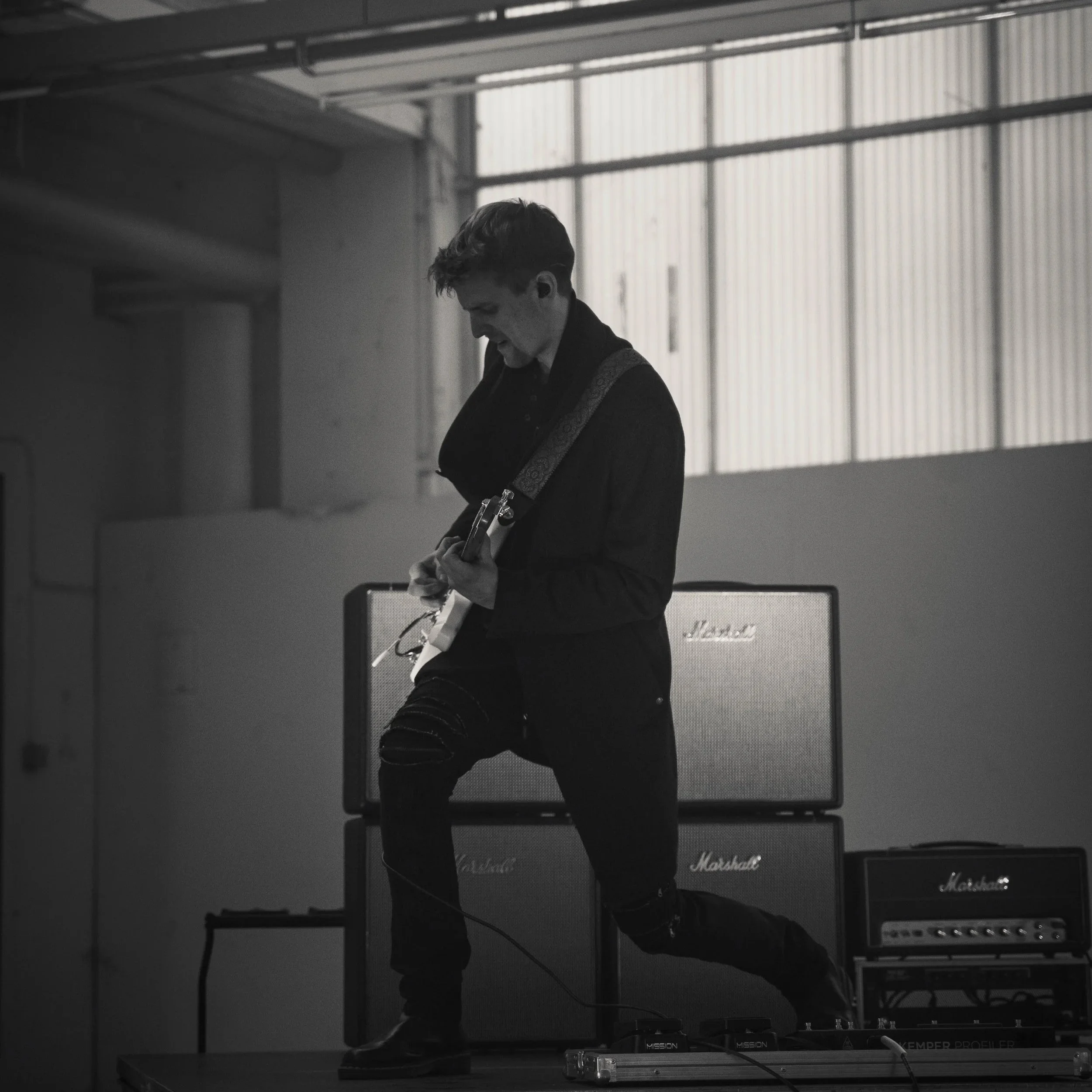 A young man playing an electric guitar on a stage in front of Marshall amplifiers, in a warehouse-like setting with large windows.