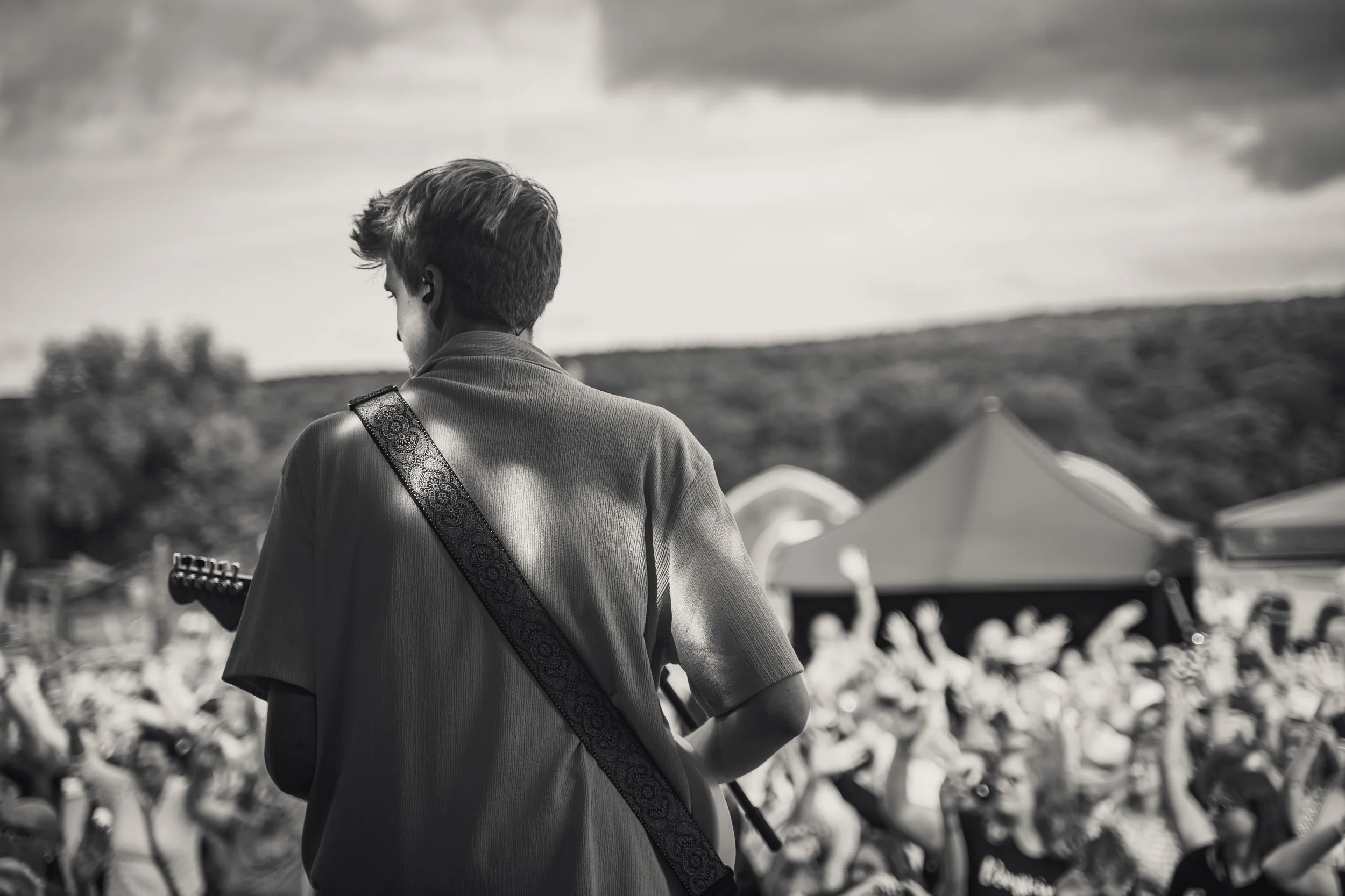 A person with short hair playing an electric guitar at an outdoor concert, with a crowd of people and tents visible in the background.