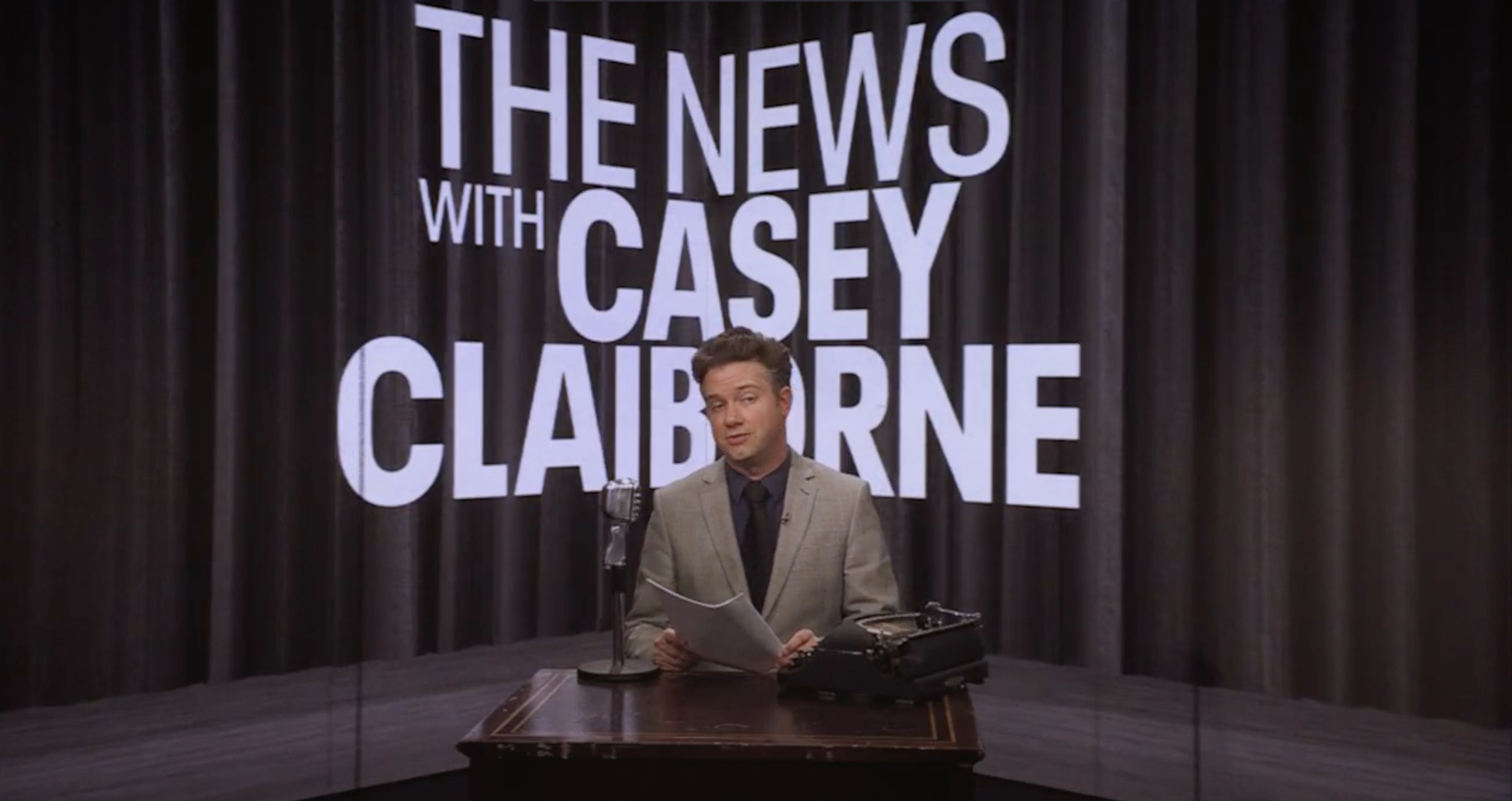 A man in a beige suit sits at a wooden desk with a vintage typewriter and microphone, in front of a black curtain with large white text that reads 'THE NEWS WITH CASEY CLAIBORNE'.
