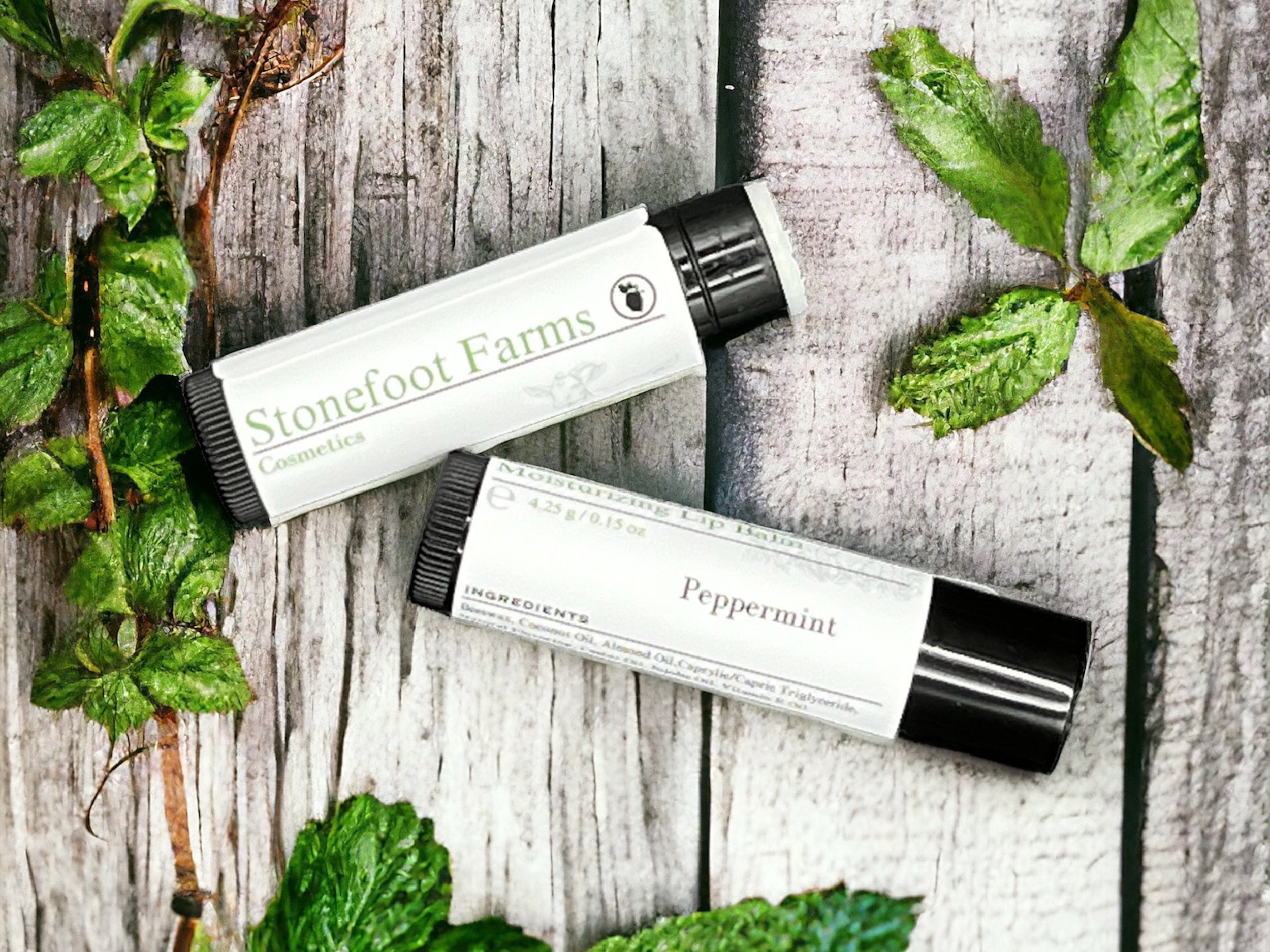 Two tubes of Stonefoot Farms Peppermint lip balm among vibrant green peppermint leaves on a textured wooden surface