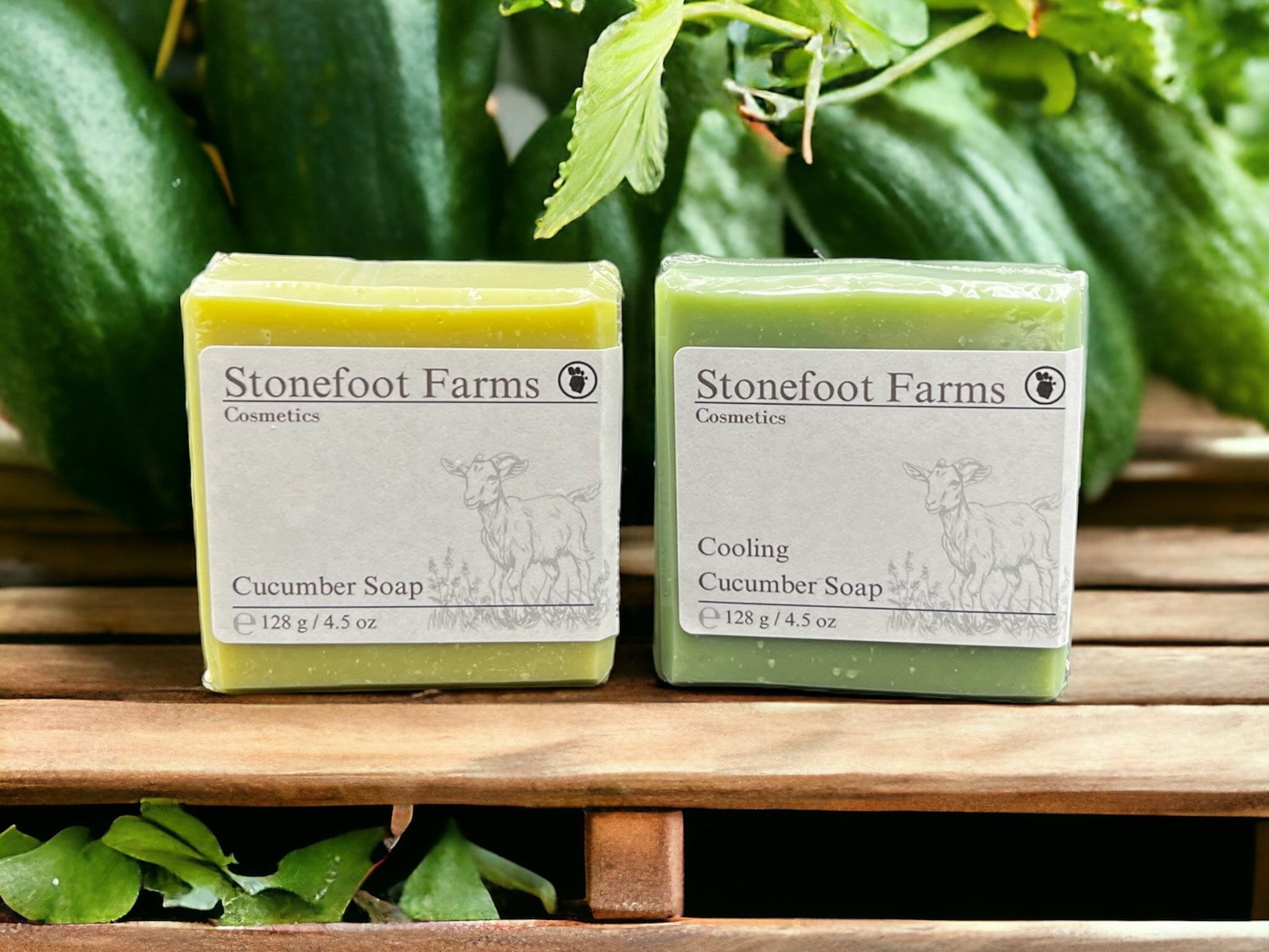 Two bars of Stonefoot Farms cucumber soap, one labeled 'Cucumber Soap' and the other 'Cooling Cucumber Soap', set against a backdrop of fresh cucumbers and green leaves on a wooden slat shelf.