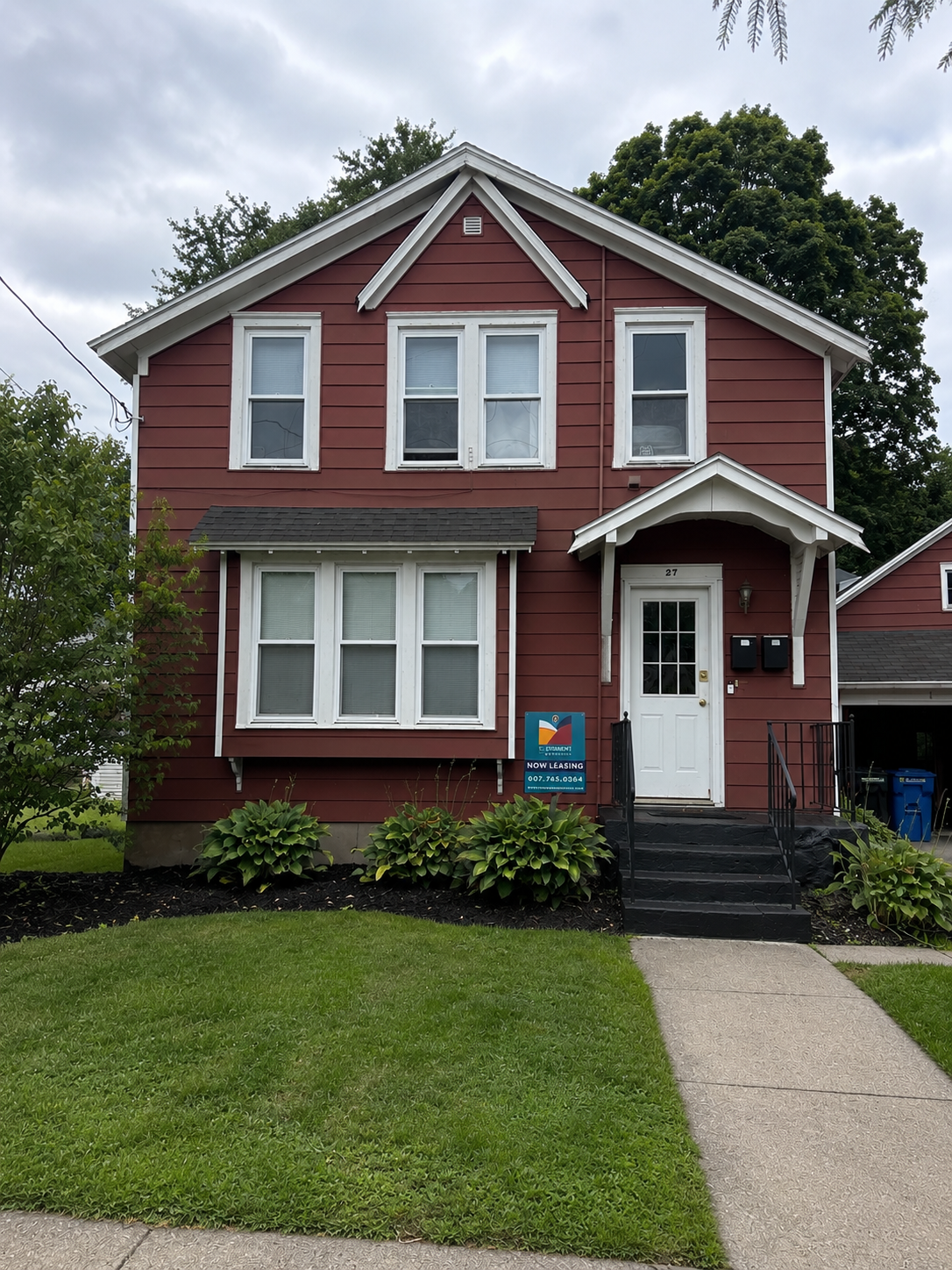 Red two-story house with white trim, front steps, and an attached garage on the right, with trees and neighboring houses in the background.