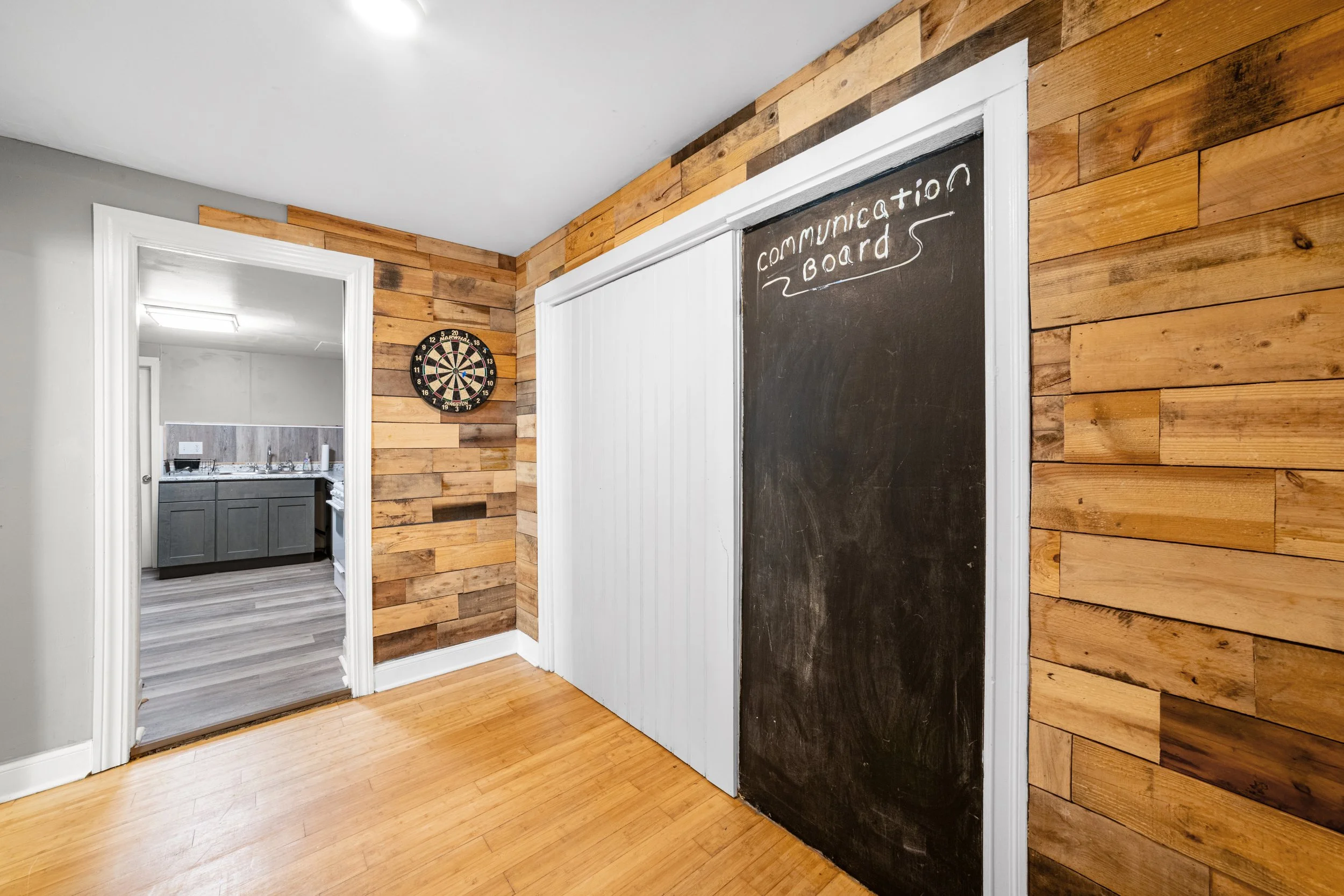 An interior room with wooden plank walls, featuring a chalkboard labeled 'Communication Board,' a white sliding door, and a door leading to a room with gray cabinetry and a dartboard on the wall.