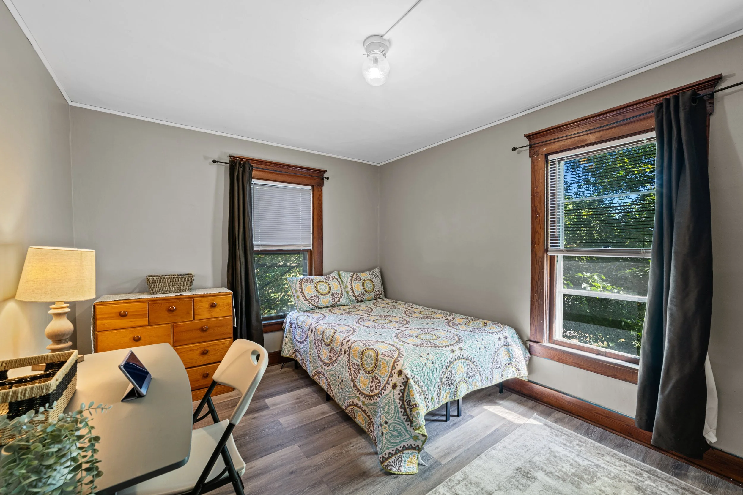 Small bedroom with a bed, a wooden dresser, a lamp, and two windows with dark curtains and wooden trim, showing a view of trees outside.