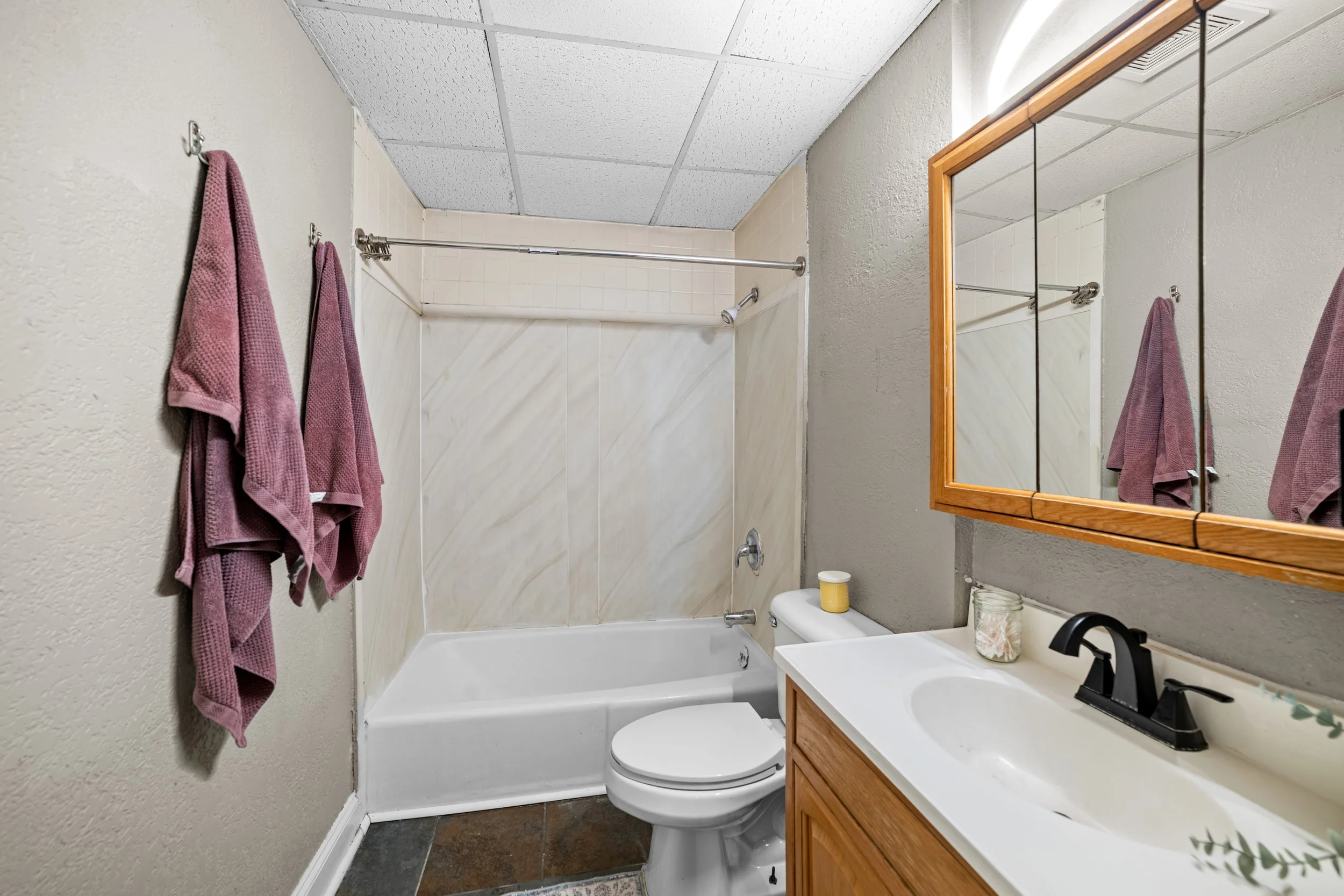 Small bathroom with a bathtub, toilet, and wooden vanity with mirror. Two pink towels hanging on the wall.