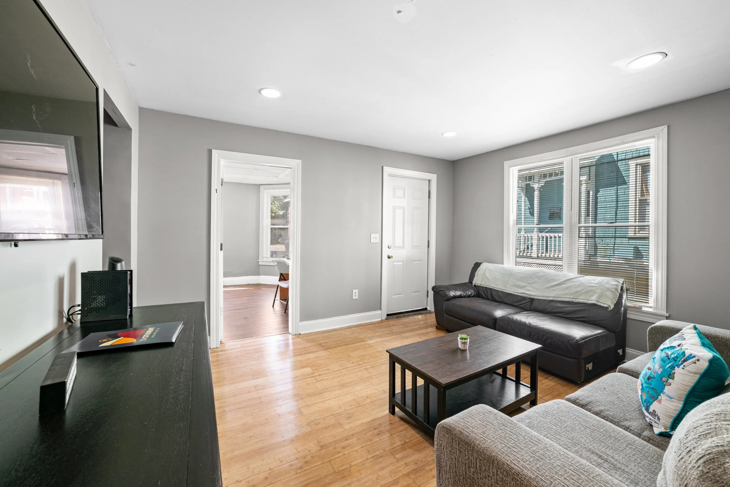 Living room with gray walls, hardwood floor, black leather couch with white throw blanket, beige sofa with colorful pillow, wooden coffee table, window with blinds, TV mounted on wall, and door leading to another room.