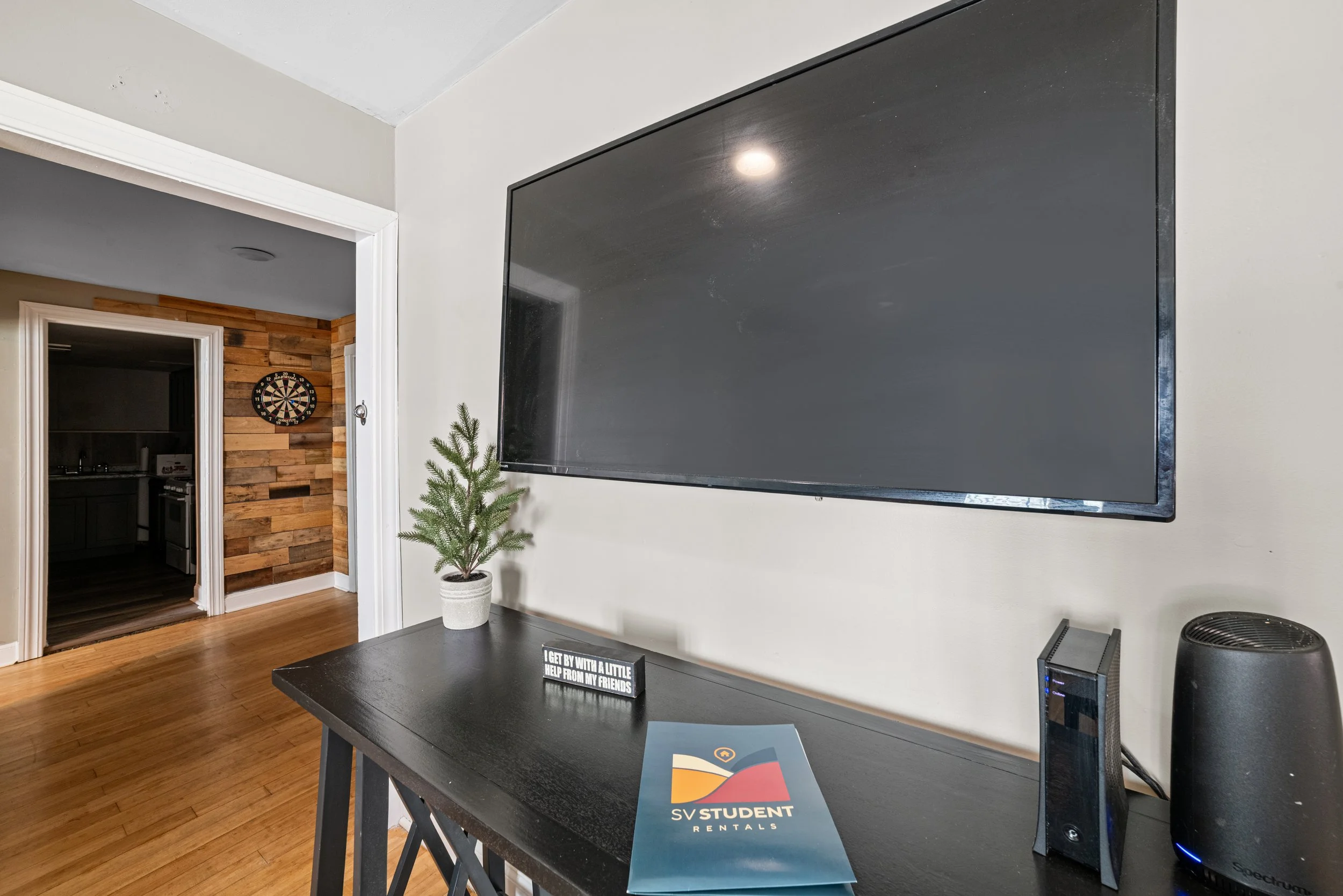 A living room with a black flat-screen TV mounted on a white wall, a small potted pine tree on a black table, and a sign that says 'GET BY WITH A LITTLE HELP FROM MY FRIENDS'. There is a router and a speaker on the table, with a wooden floor and a do