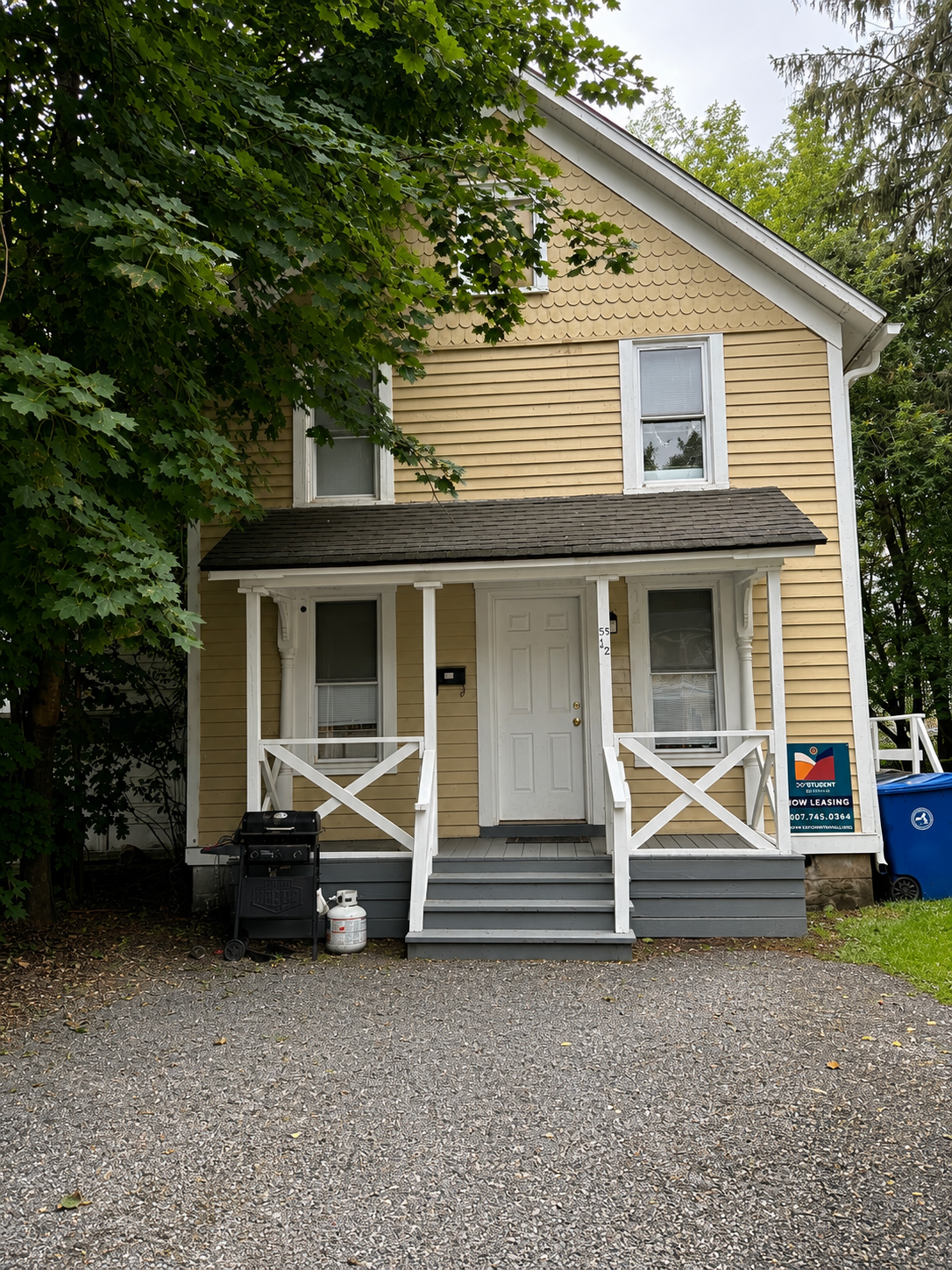 A beige two-story house with white trim, a small front porch, and a sign for student rentals. The house is surrounded by green trees and has a dirt driveway.