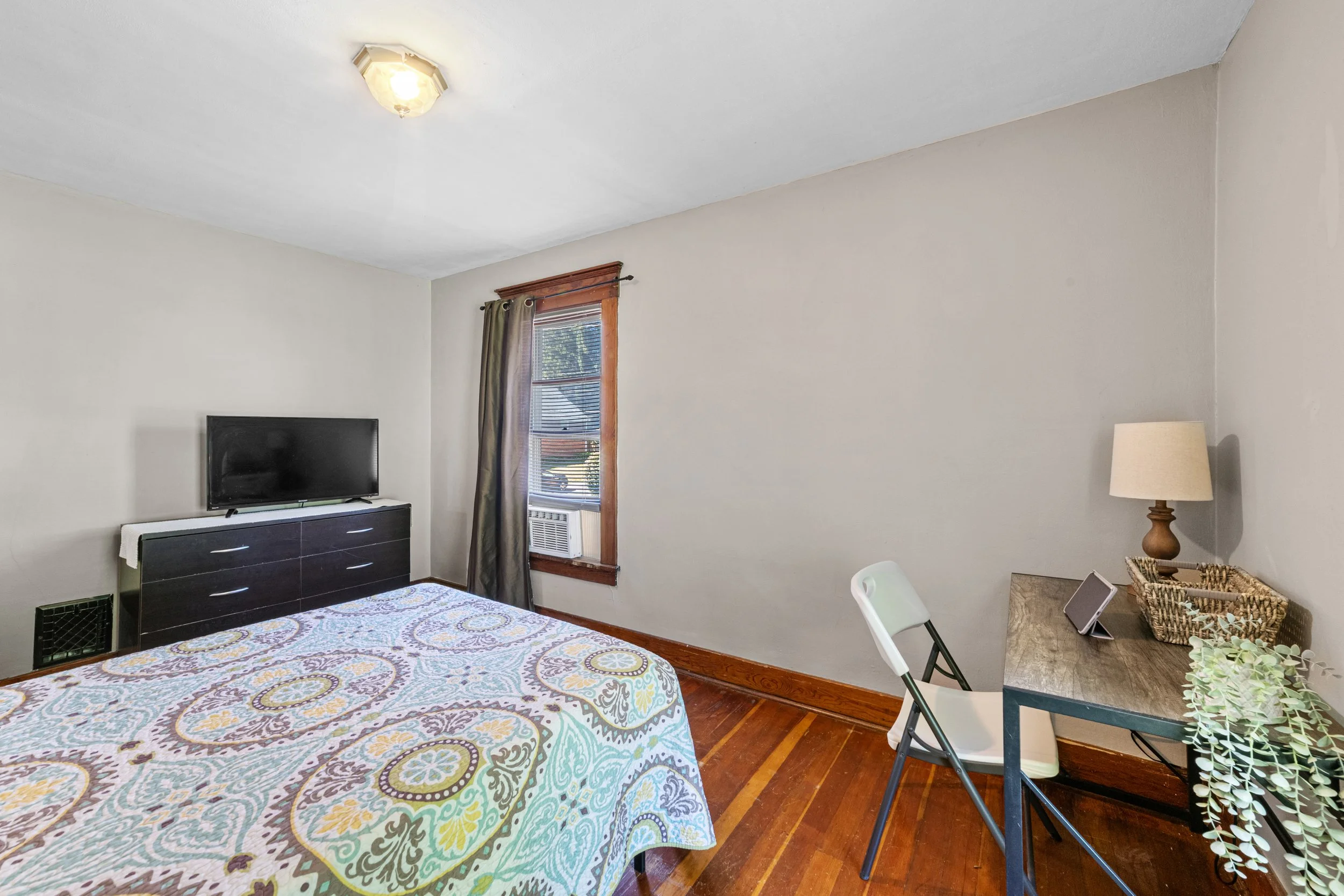 A bedroom with a bed, a TV on a dresser, a window with curtains, a small desk with a lamp, a tablet, a folding chair, and hardwood floors.