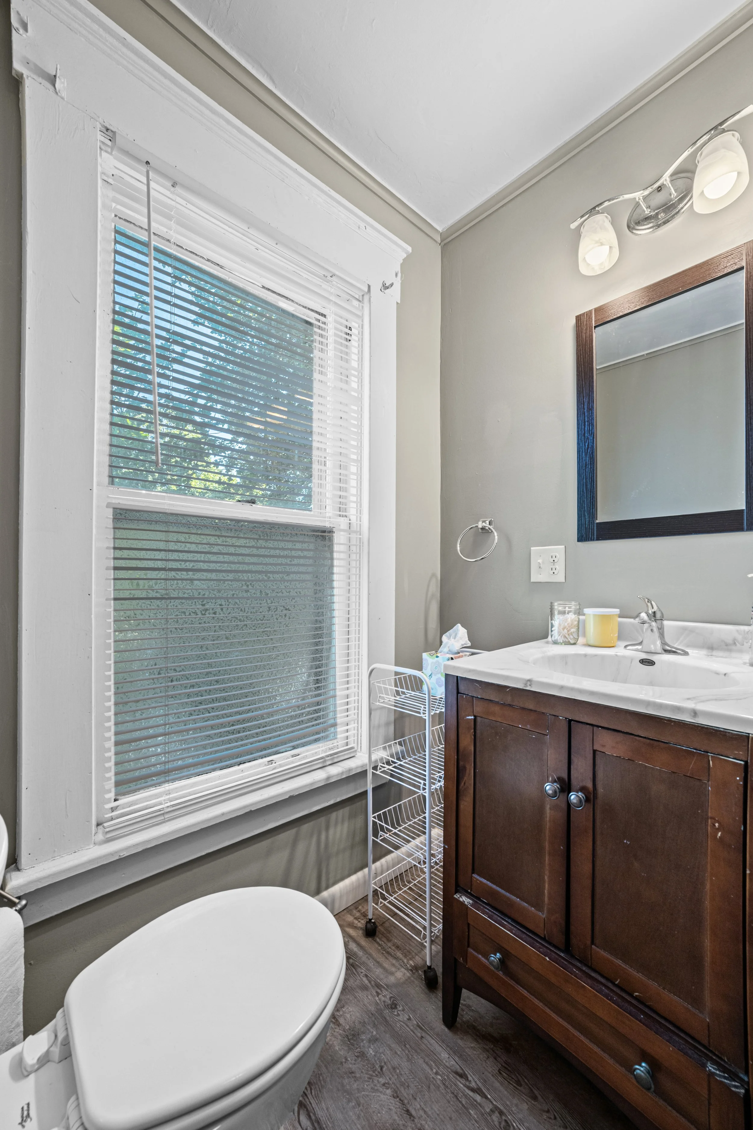 A small bathroom with a window, white blinds, a wooden vanity with a marble top, a mirror, and a toilet. There is a small white storage cart next to the vanity.
