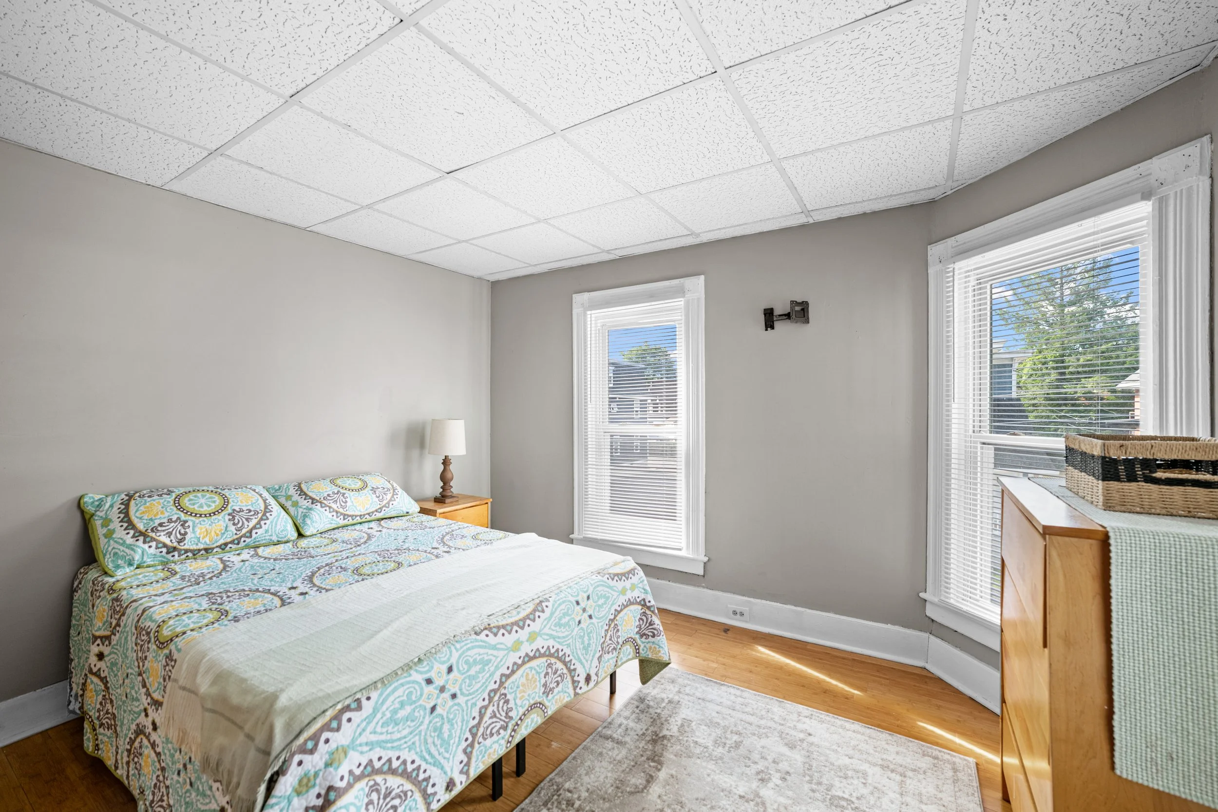 A bedroom with a bed, nightstand with lamp, and dresser. Two windows with blinds letting in sunlight. Light-colored walls, hardwood floor, and white ceiling tiles.