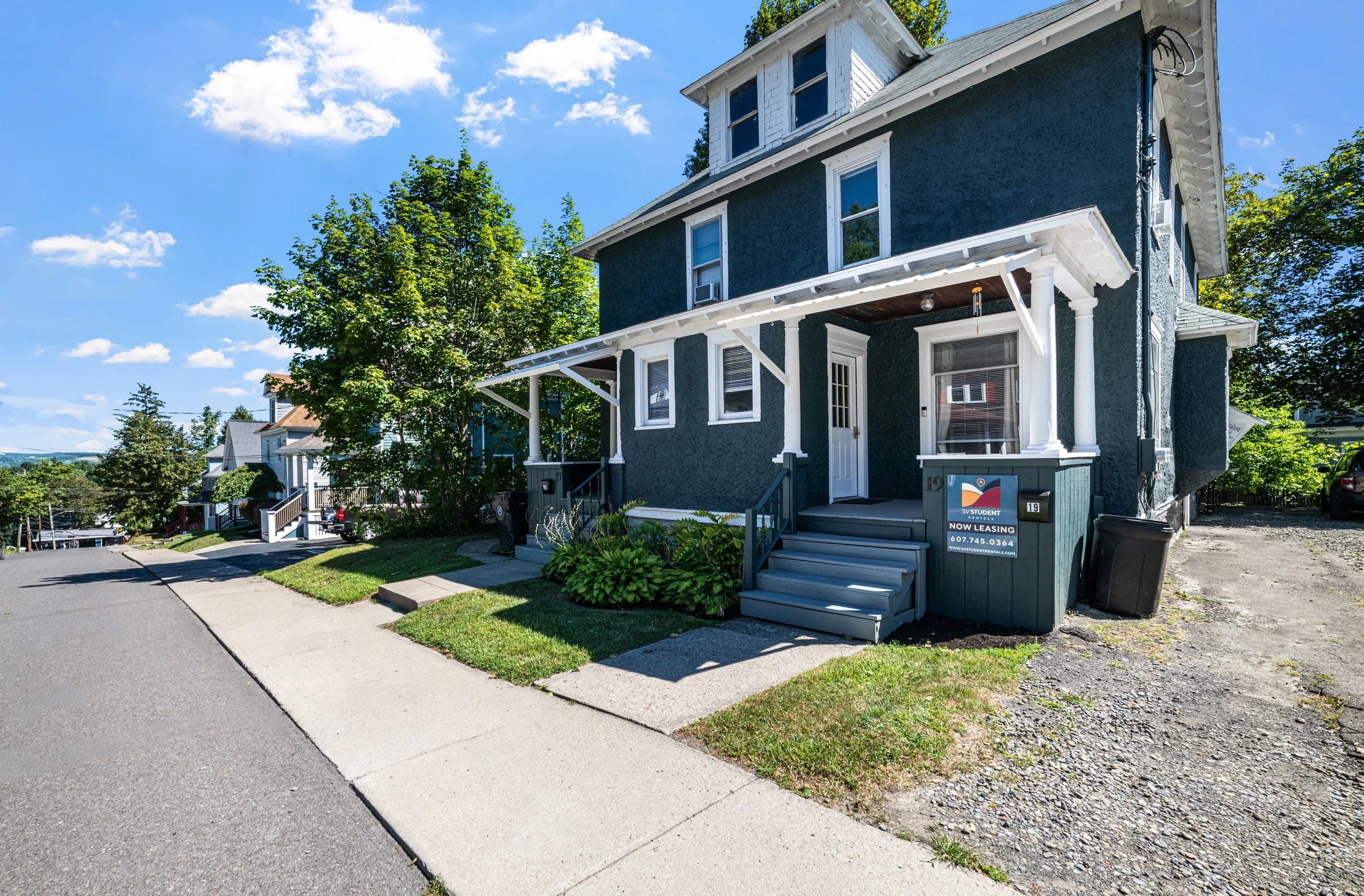 A blue, three-story house on a suburban street with a sidewalk, steps leading to the front porch, green bushes, and a 'Now Leasing' sign in the yard.