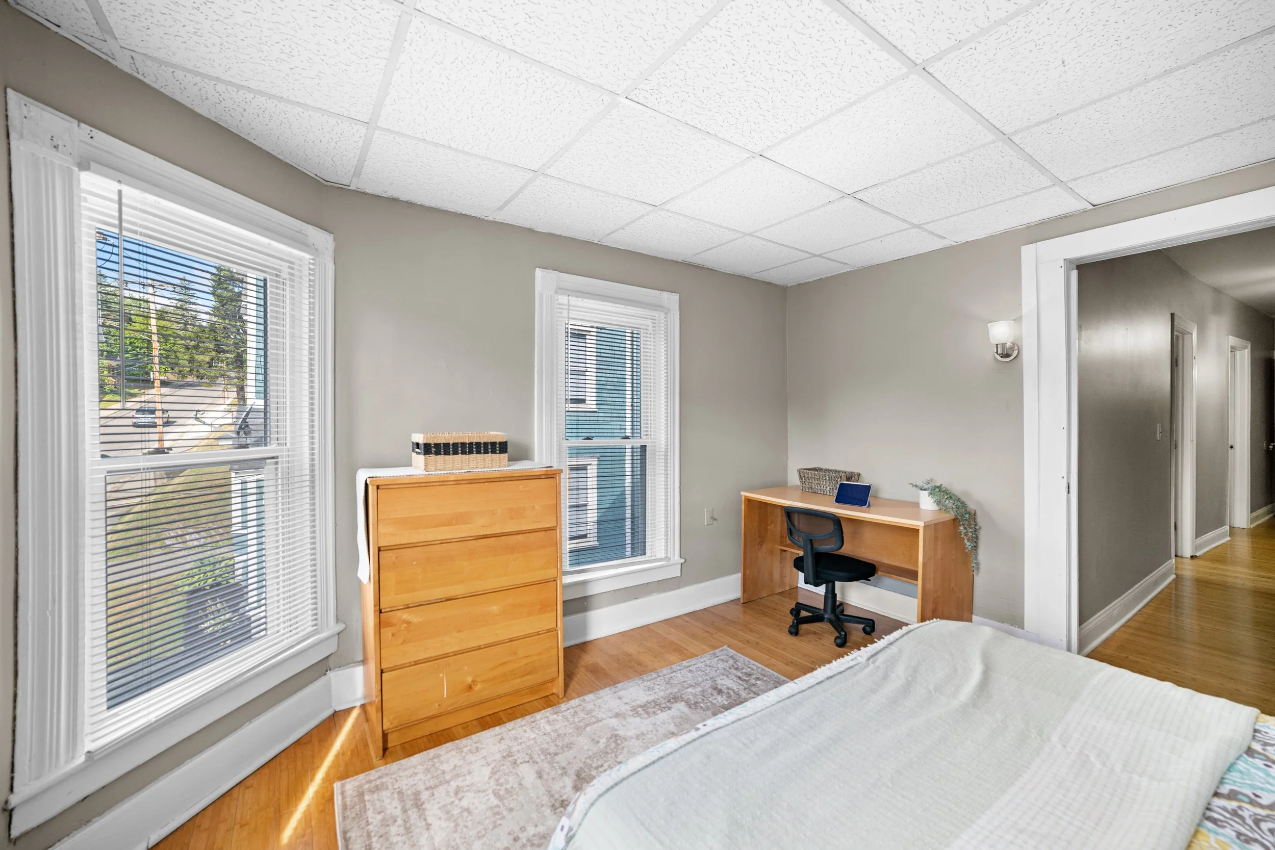A bedroom with two large windows, a wooden dresser, a desk with a chair, and a bed with a white blanket, hardwood floors, and light-colored walls.