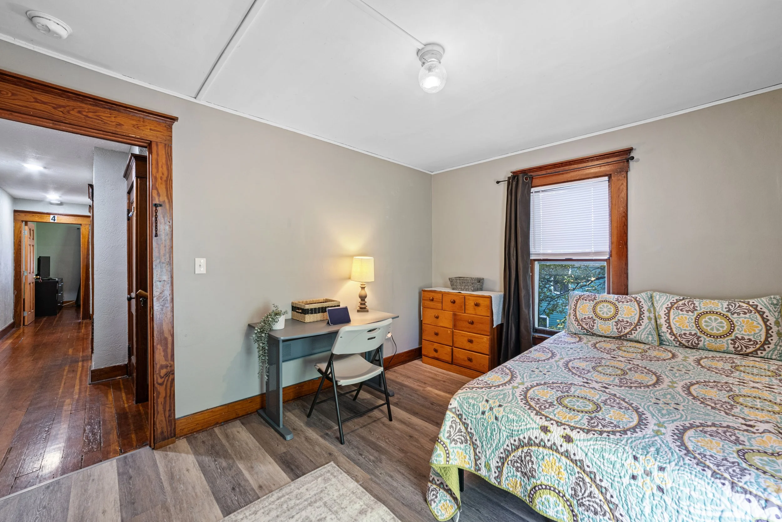 A bedroom with a bed, a dresser, a window with blinds and curtains, a desk with a chair and a lamp, and a hallway leading to other rooms.