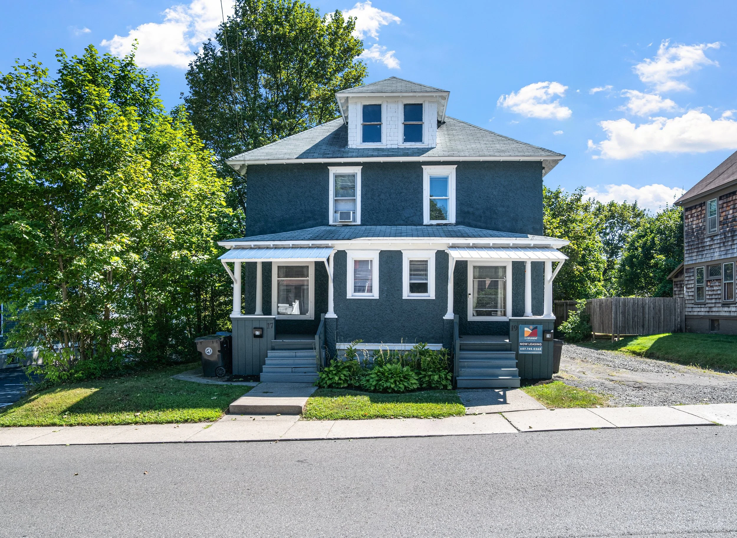 A three-story house painted dark blue with white trim and a small front porch, situated on a residential street with trees and neighboring houses visible.