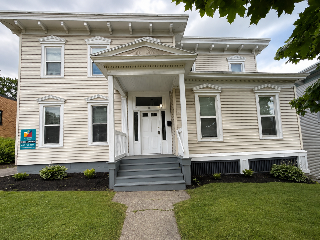 A beige three-story house with white trim, a small front porch with gray stairs, and multiple windows, some with curtains. There are trees and a grassy yard, with an uneven driveway on the left and a stone staircase on the right.