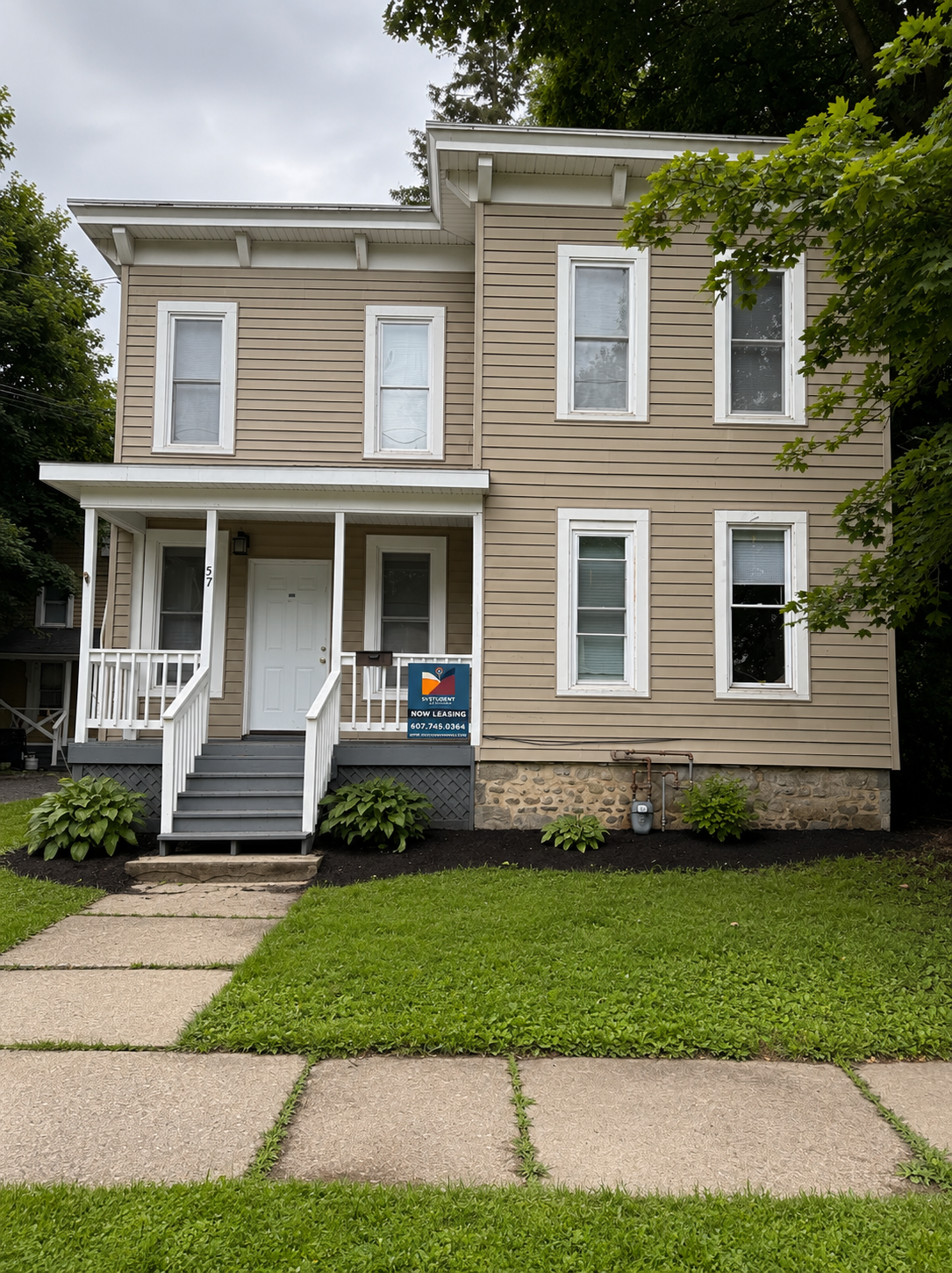 Front view of a two-story beige house with white trim and a small porch with steps leading up to it, surrounded by trees and greenery.