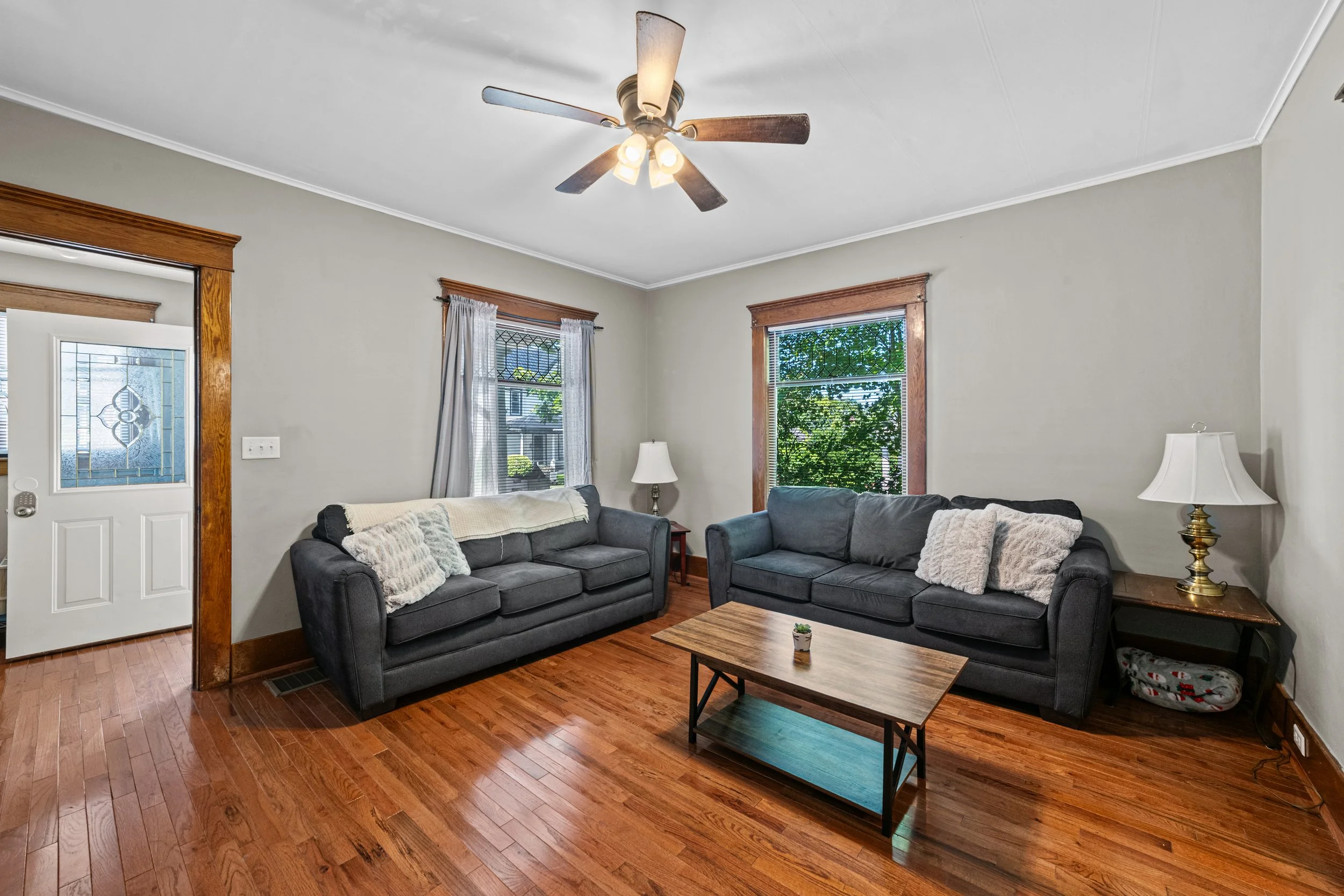 Living room with two dark gray sofas, a small wooden coffee table, wooden trim around windows and door, hardwood floor, ceiling fan with lights, and two table lamps.