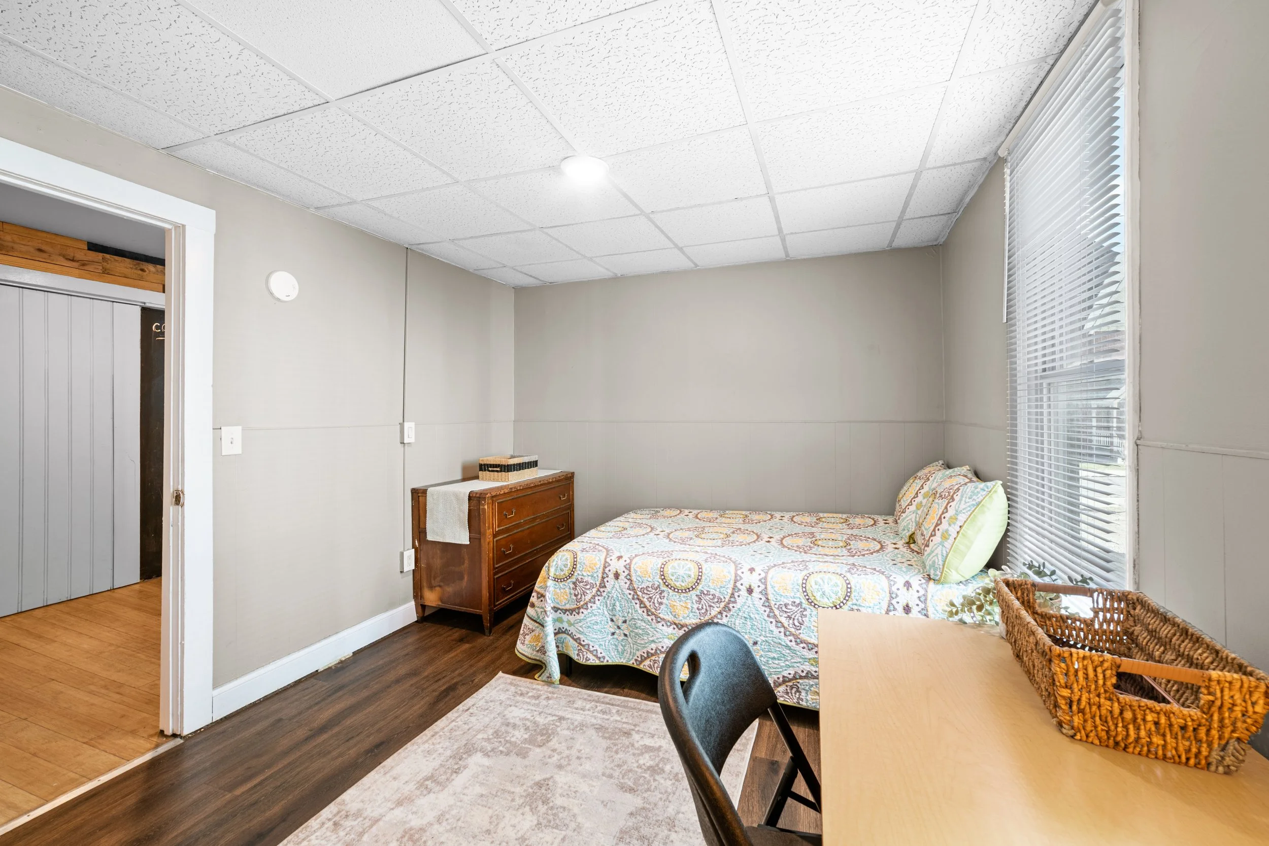 A simple bedroom with a single bed covered with a patterned quilt, a window with blinds, a wooden dresser, and a small table with a wicker basket on top.