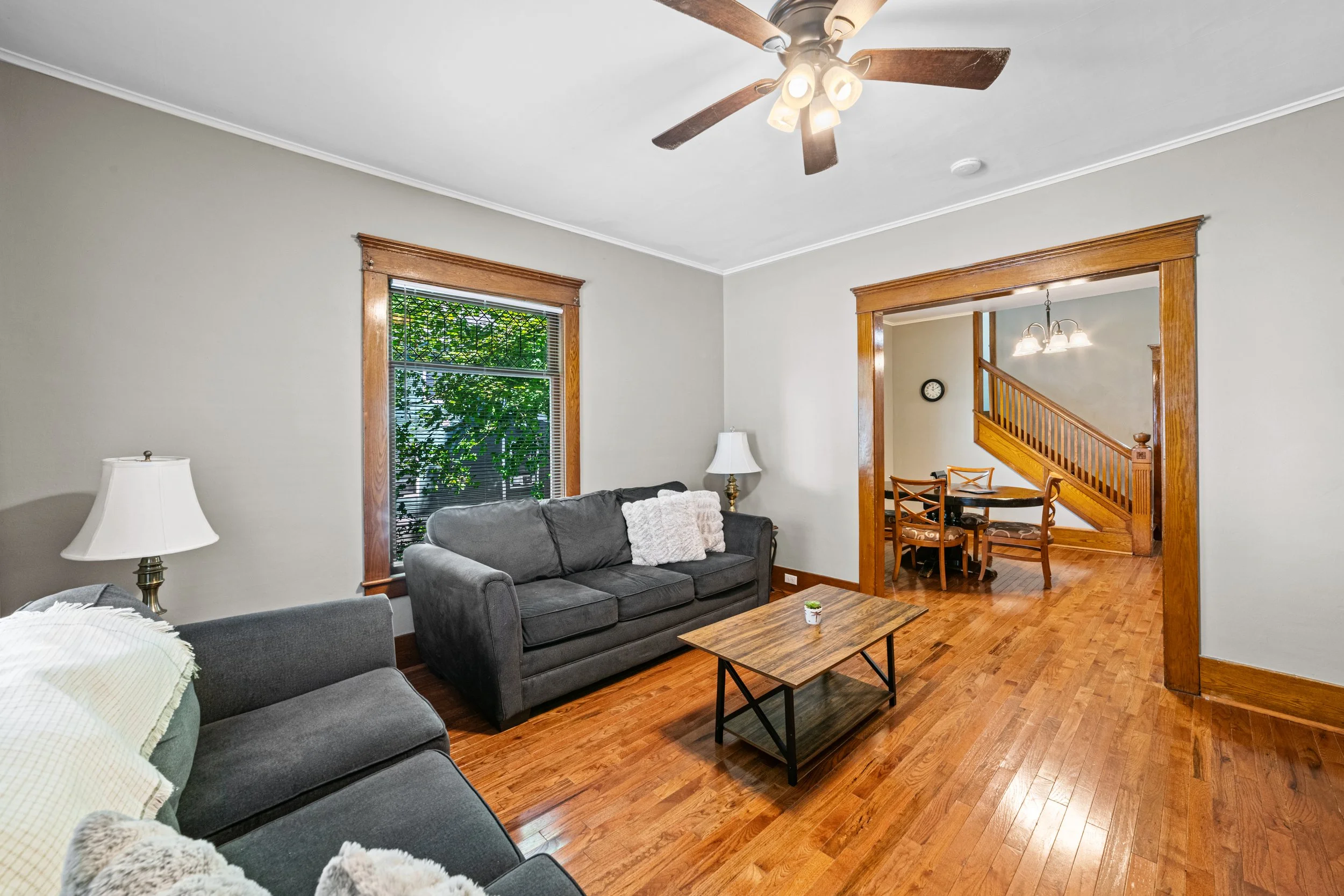 Living room with hardwood floors, gray sofas with blankets, wooden window frames, a ceiling fan with lights, and an opening leading to a dining area with a wooden staircase and chairs.