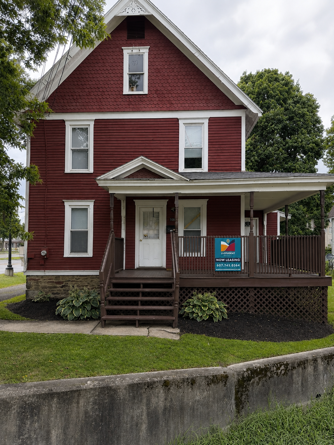 A red three-story house with a front porch, white trim, and a sign advertising student rentals. The house is surrounded by green grass and trees.