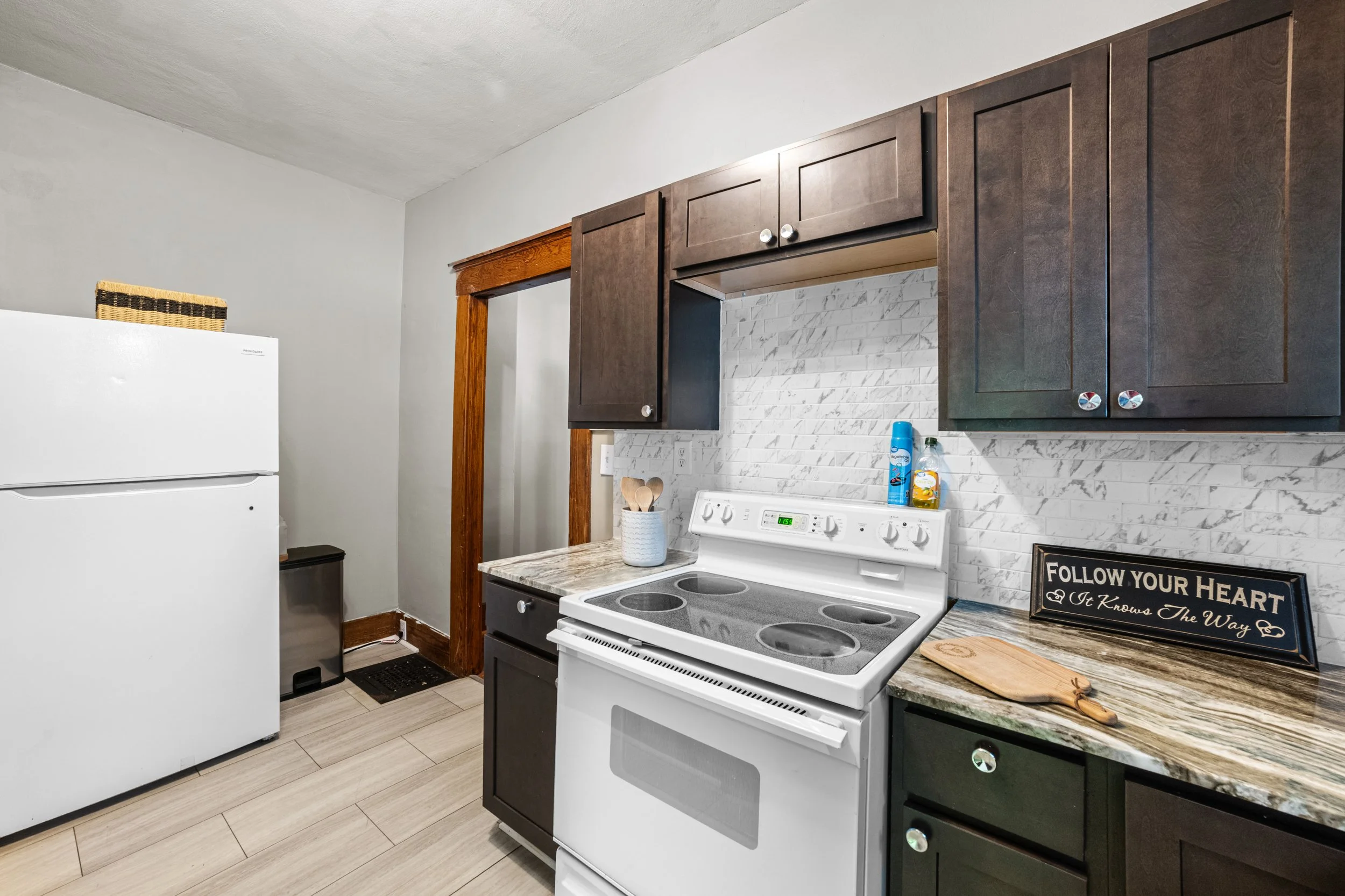 Kitchen with white refrigerator, white stove, dark wood cabinets, marble backsplash, and countertop. There is a sign that says "Follow Your Heart" on the counter.